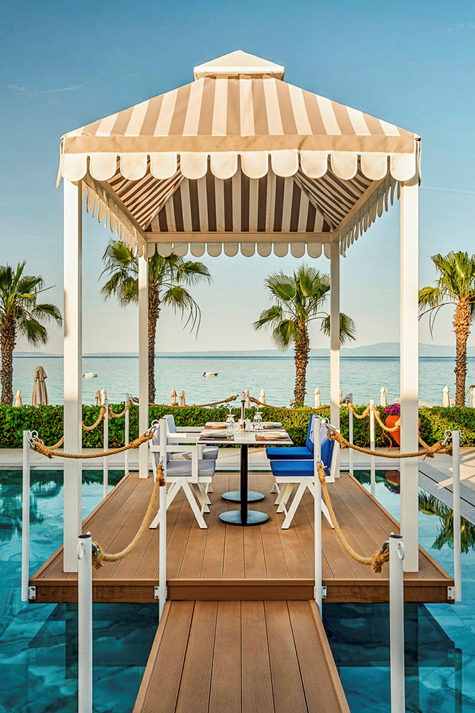 En paviljong med ett matbord och stolar på en brygga vid en pool. I bakgrunden syns palmer och havet. (AI-genererad text)