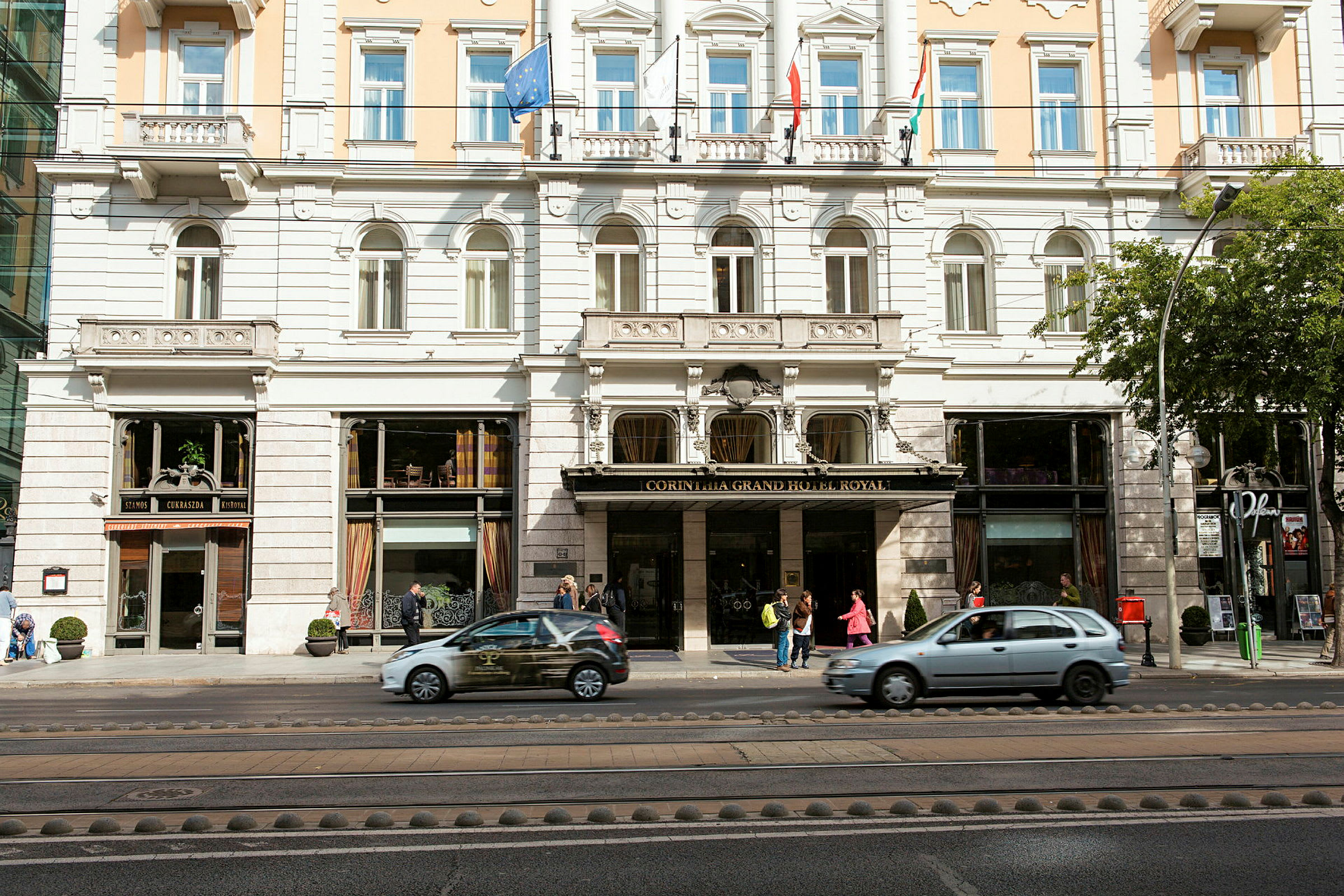 Fasad av ett hotell med flera våningar, invändigt synliga fönster och entréområdet med människor och förbipasserande bilar. (AI-genererad text)