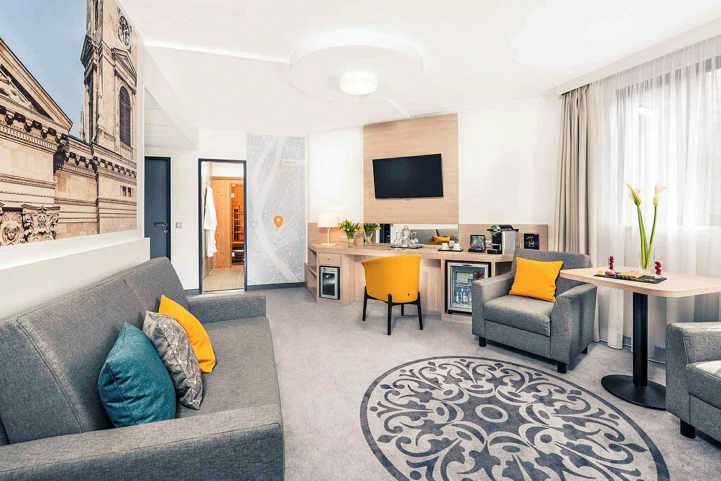 Ett hotellrum med sittgrupp, skrivbord, och en stor väggbild av en byggnadsfasad. (AI-genererad text)