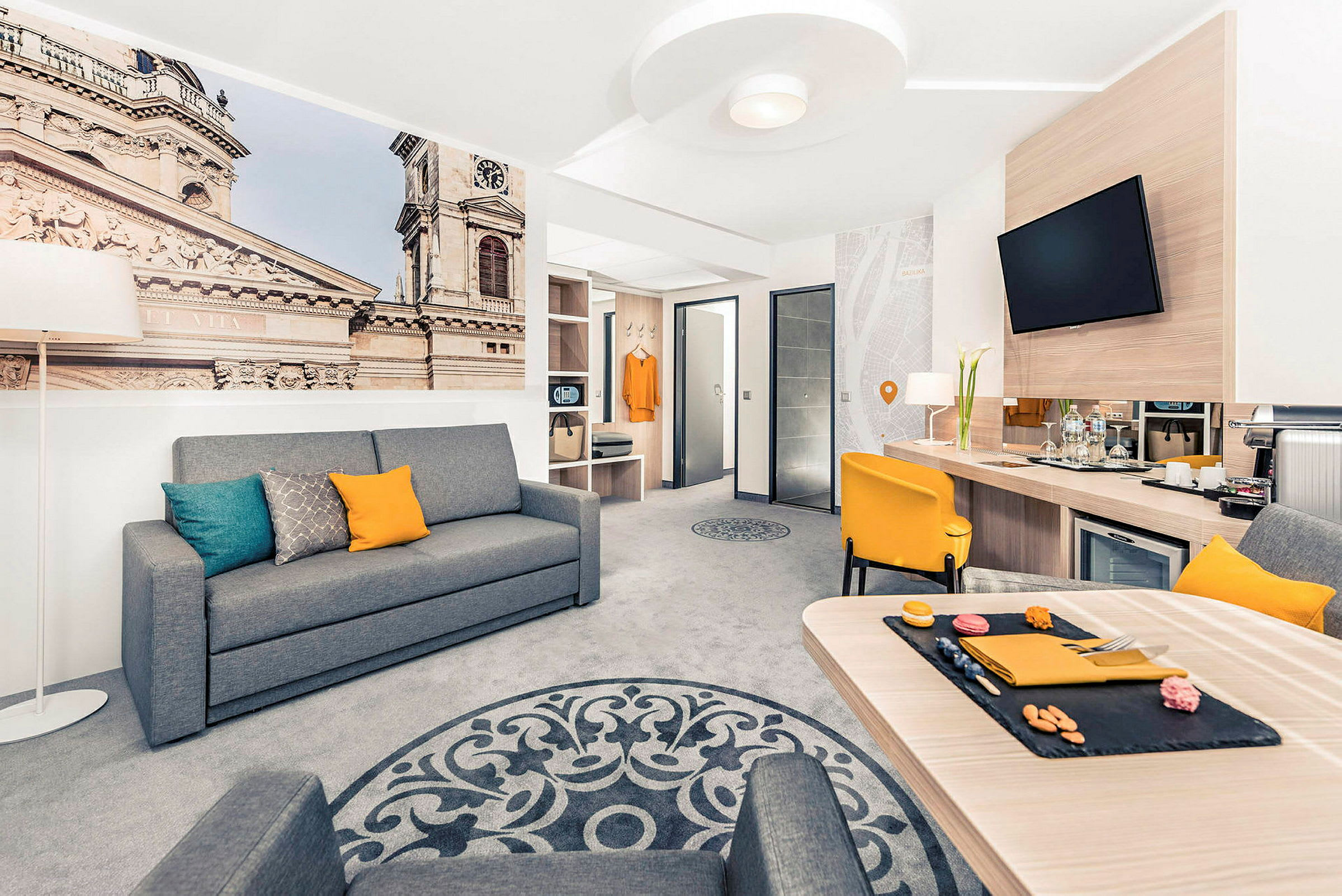 Interiör av ett modernt hotellrum med en grå soffa, gul stol och ett bord, samt en stor väggbild som visar en byggnad. (AI-genererad text)