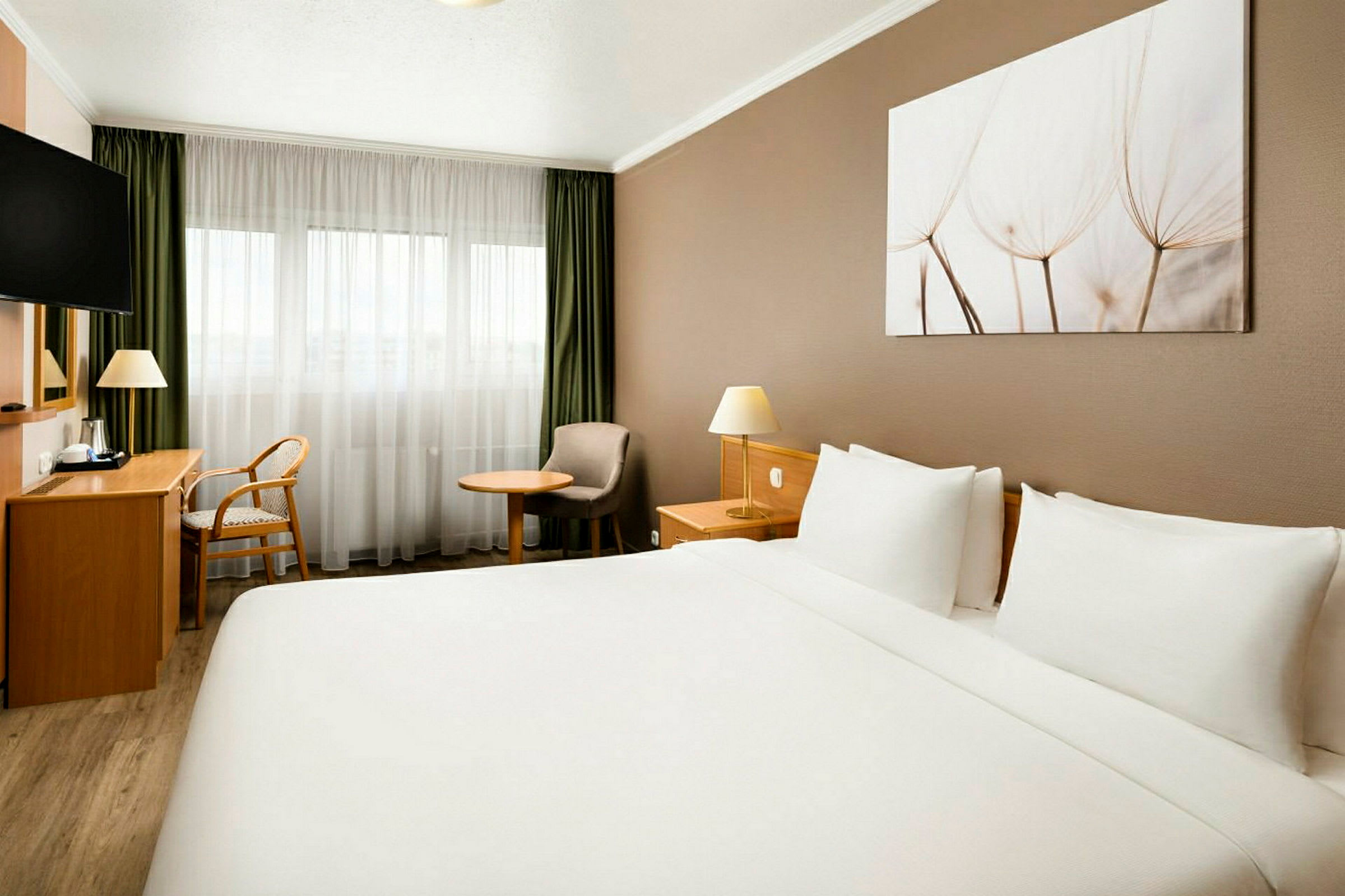 Ett hotellrum med en stor dubbelsäng, skrivbord, stol, fåtölj och fönster med ljusa gardiner. (AI-genererad text)