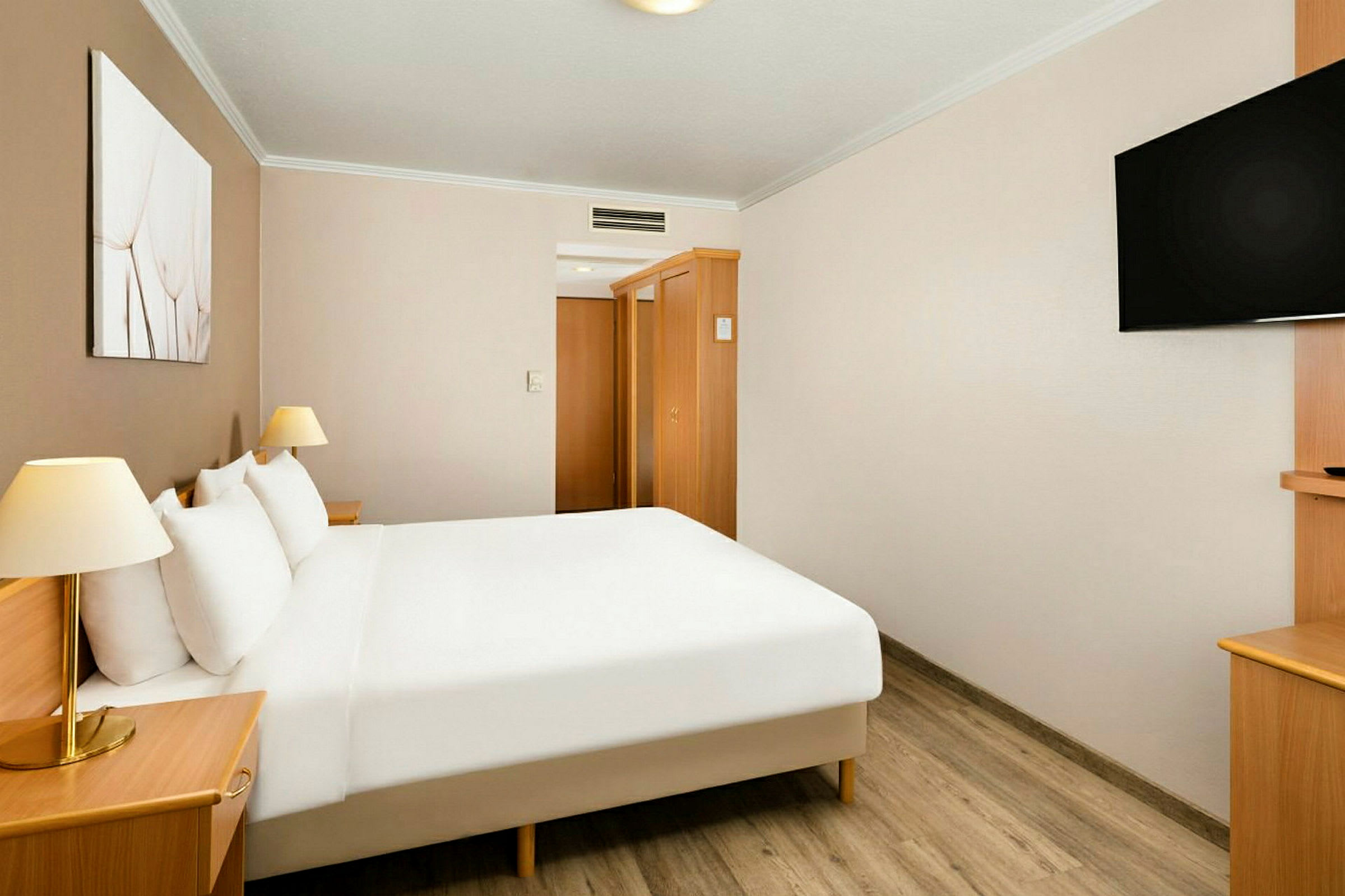 Ett hotellrum med en dubbelsäng, vita lakan, nattduksbord med lampor, väggmonterad TV och trädetaljer på möblerna. (AI-genererad text)