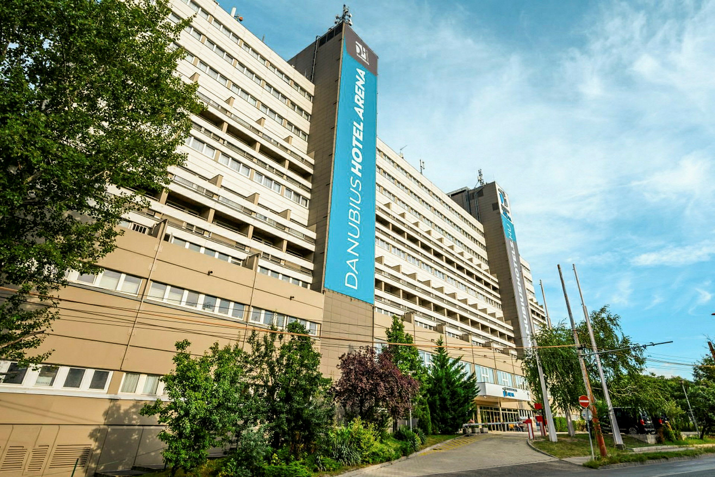 En stor byggnad med hotellnamnet 'Danubius Hotel Arena' synligt på en skylt. Träd och växtlighet omger området och en blå himmel kan ses ovanför. (AI-genererad text)