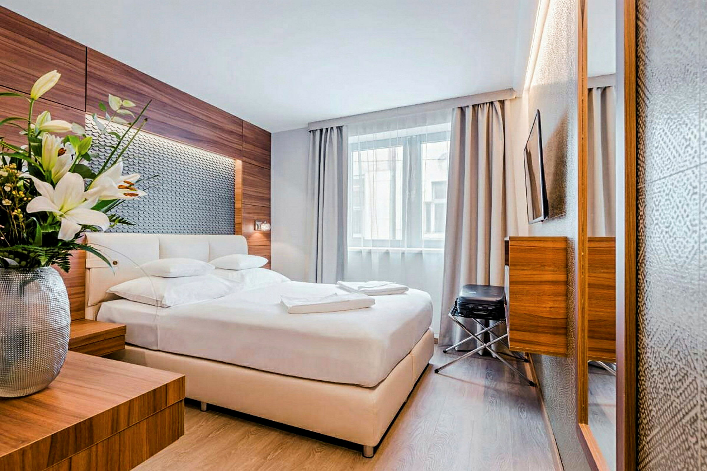 Ett modernt hotellrum med en dubbelsäng, ett stort fönster med gardiner och fräscht inredning med trädetaljer. En vas med blommor står på ett nattduksbord. (AI-genererad text)