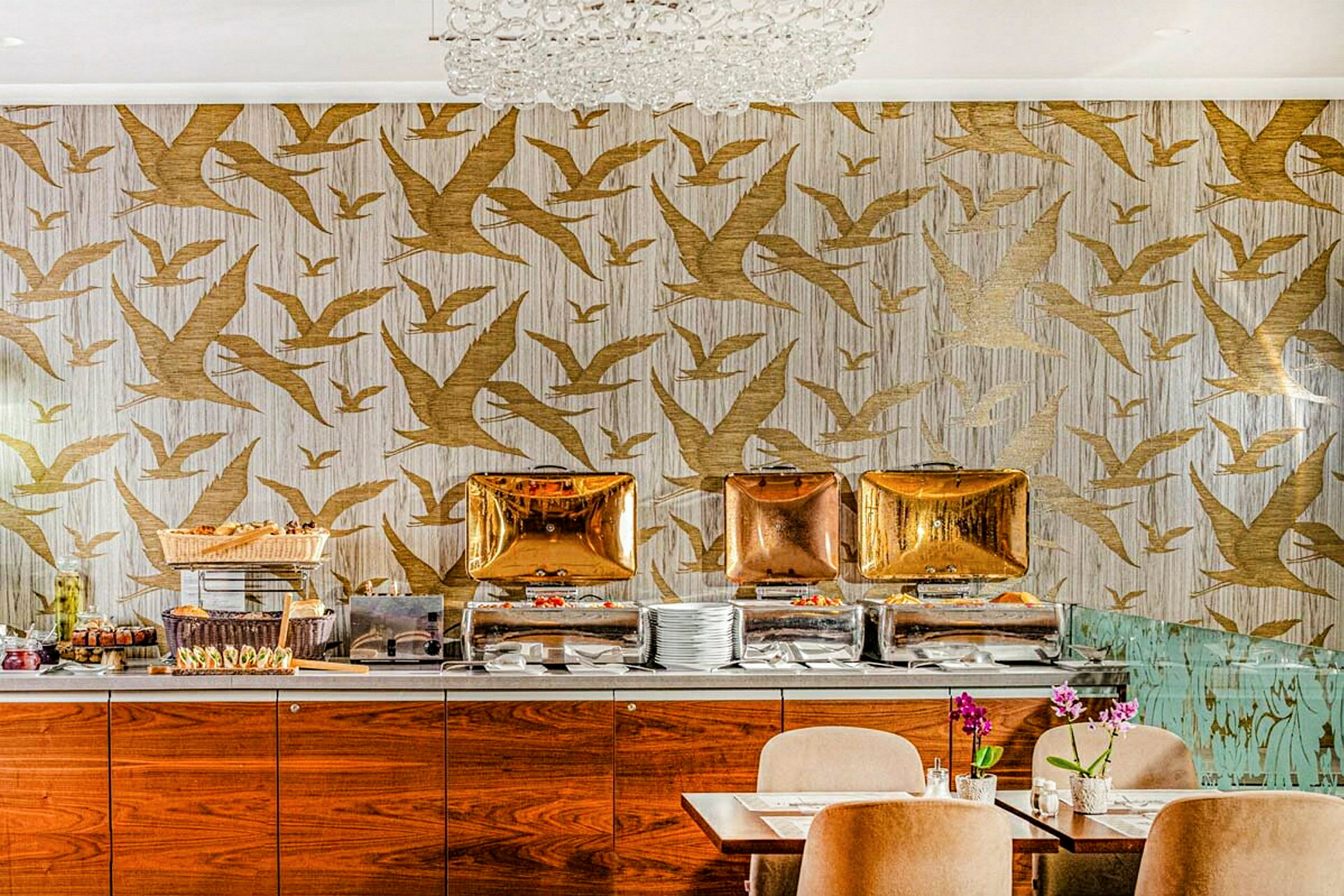 En buffé med mat står framdukad framför en tapet med fågelmotiv i guld. Framför står matsalsbord med stolar. (AI-genererad text)