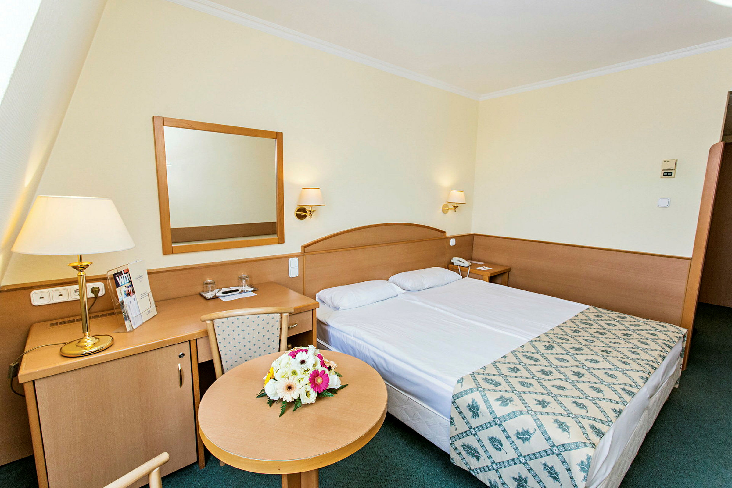 Hotellrum med dubbelsäng, skrivbord och blommor på ett runt bord. (AI-genererad text)
