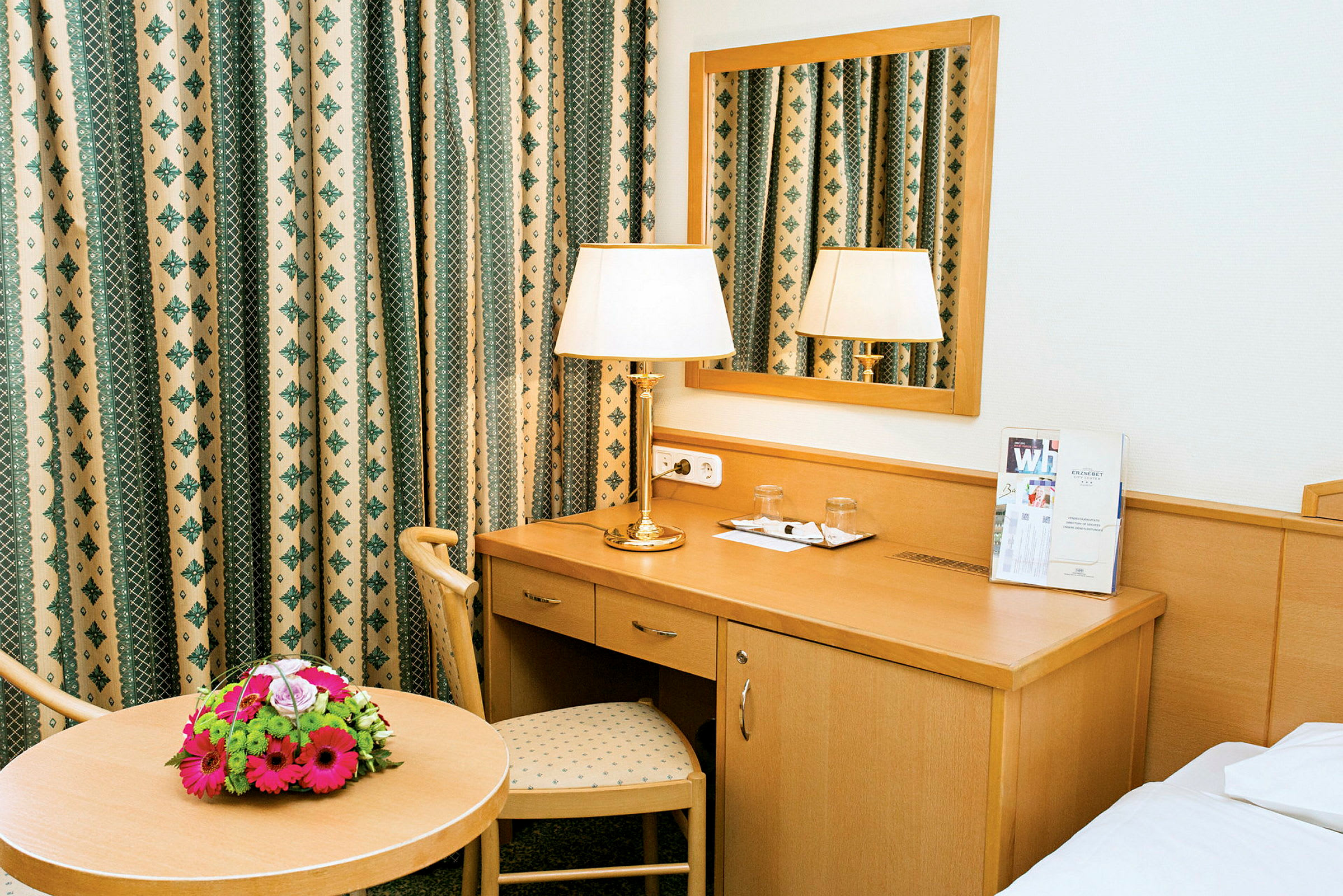 Ett hotellrum med ett skrivbord, spegel, bord med blombukett och lampa. (AI-genererad text)
