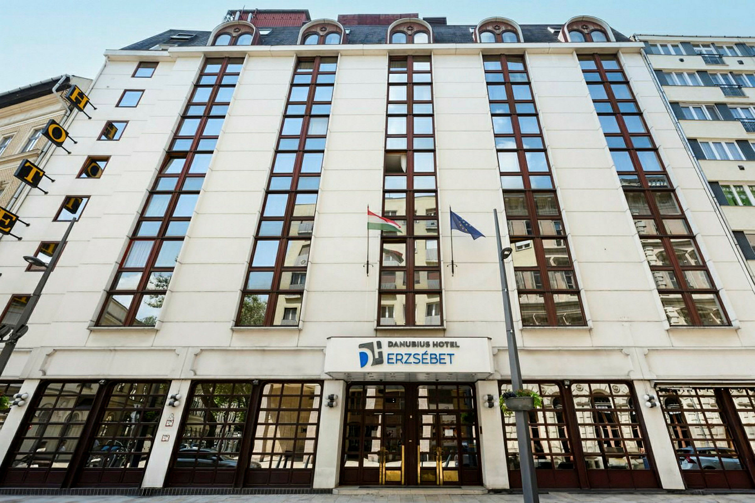 Framsidan av Danubius Hotel Erzsébet med glasfönster och flaggor. (AI-genererad text)