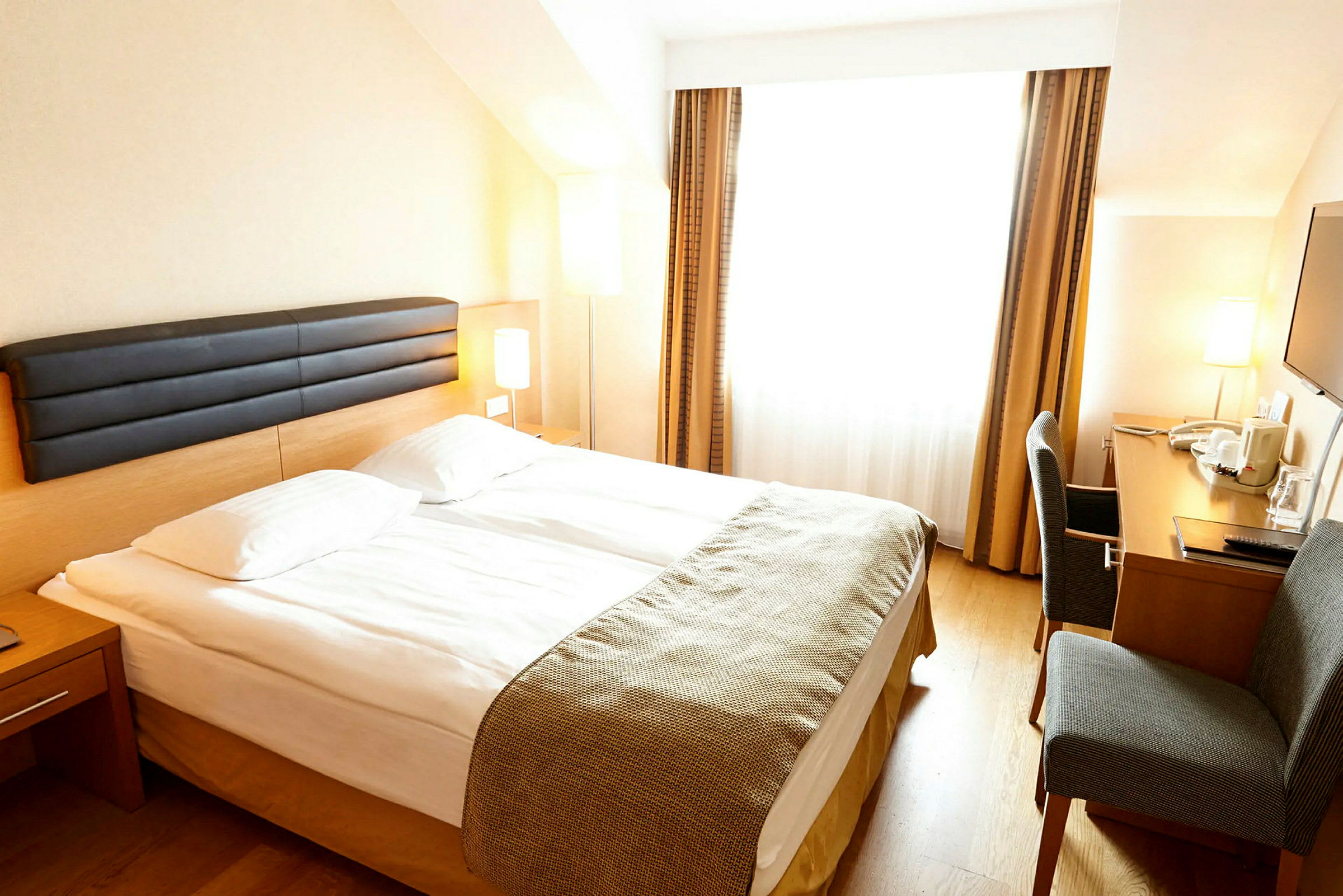 Hotellrum med en dubbelsäng, ett skrivbord och en stol. Rummet har stora fönster med gardiner. (AI-genererad text)