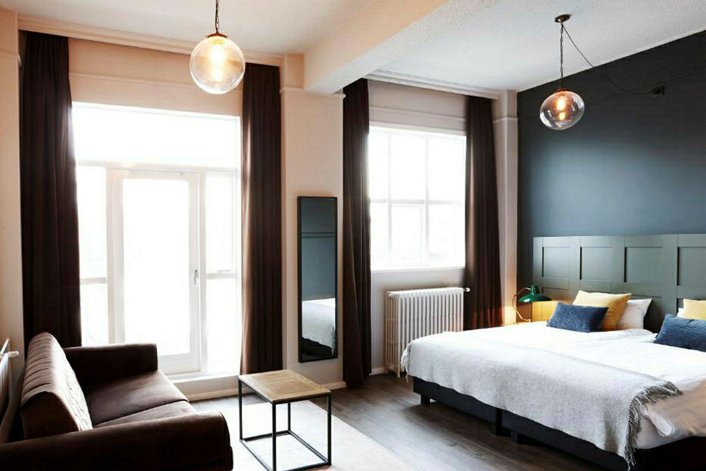Ett hotellrum med en dubbelsäng, en soffa och ett litet soffbord samt stora fönster med gardiner. (AI-genererad text)