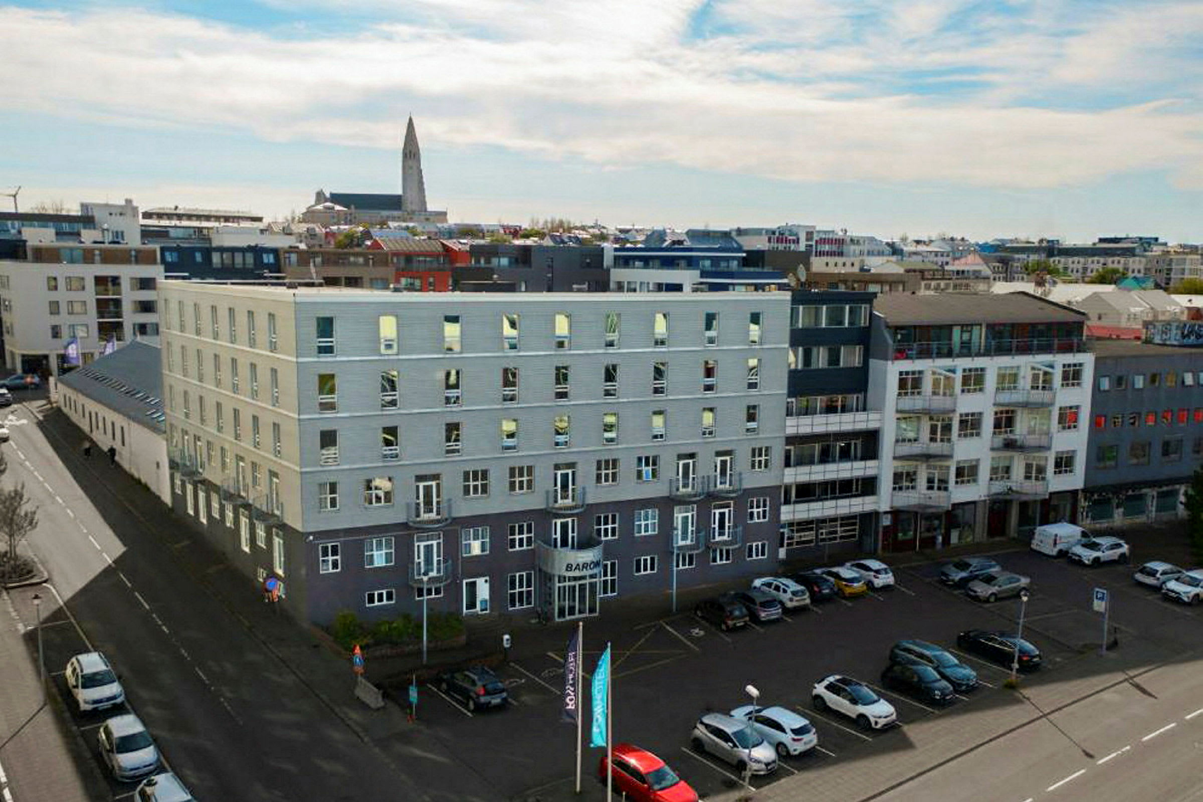 Flera moderna byggnader omger ett öppet parkeringsområde, med bilar parkerade på marken och en kyrka synlig i bakgrunden. (AI-genererad text)