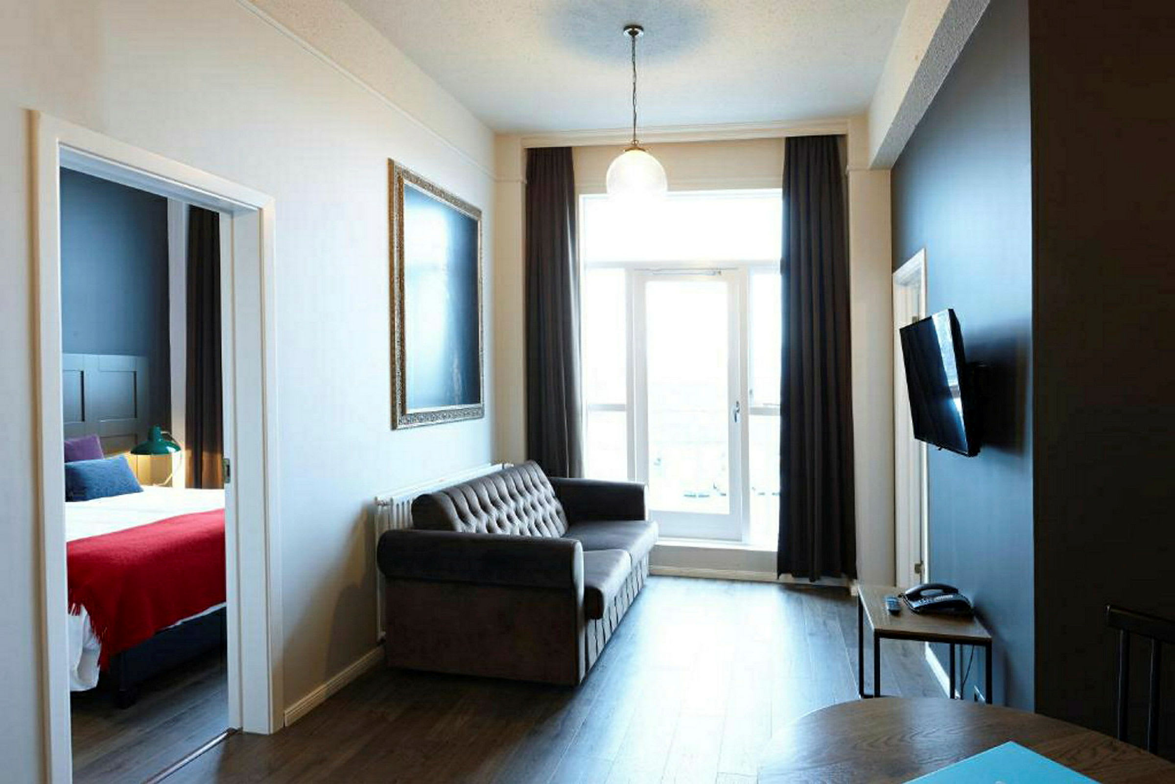 Ett vardagsrum med en soffa, en väggmonterad TV, ett litet bord och stor fönsterdörr som leder till en balkong. Ett sovrum visas i dörröppningen till vänster. (AI-genererad text)