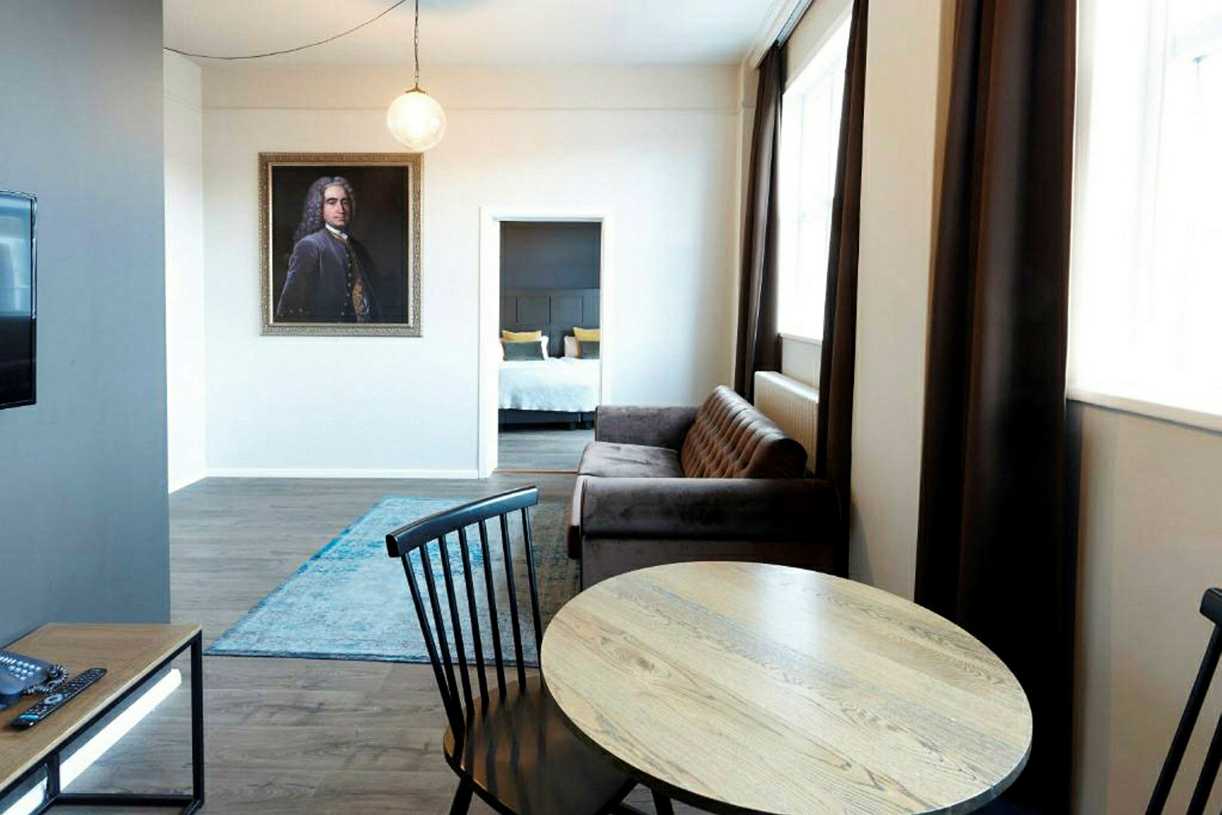 Ett vardagsrum med bord och stolar framför en soffa, en matta på golvet och en passage till ett sovrum i bakgrunden. (AI-genererad text)
