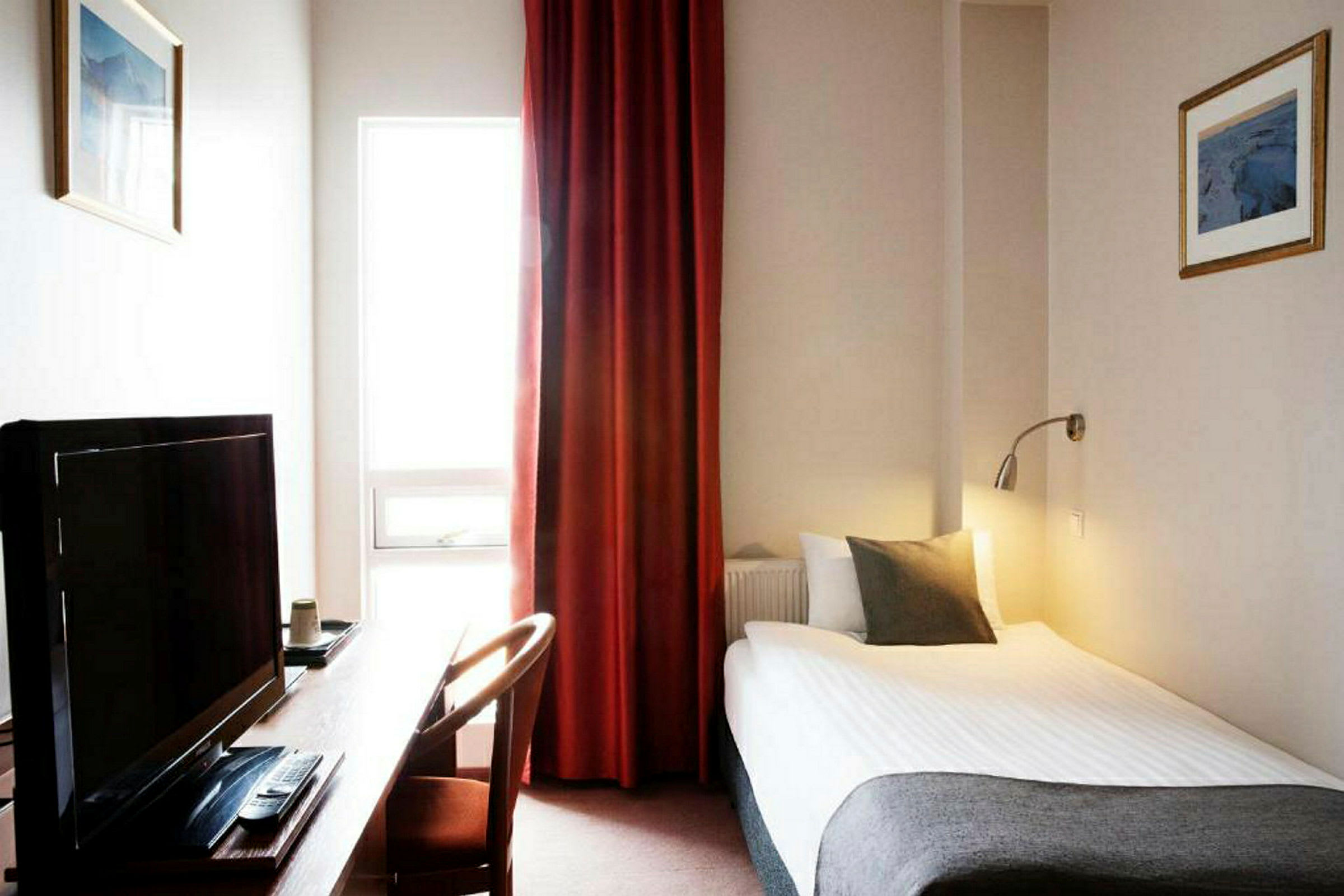 Ett hotellrum med en enkelsäng, skrivbord, TV och fönster med röda gardiner. (AI-genererad text)