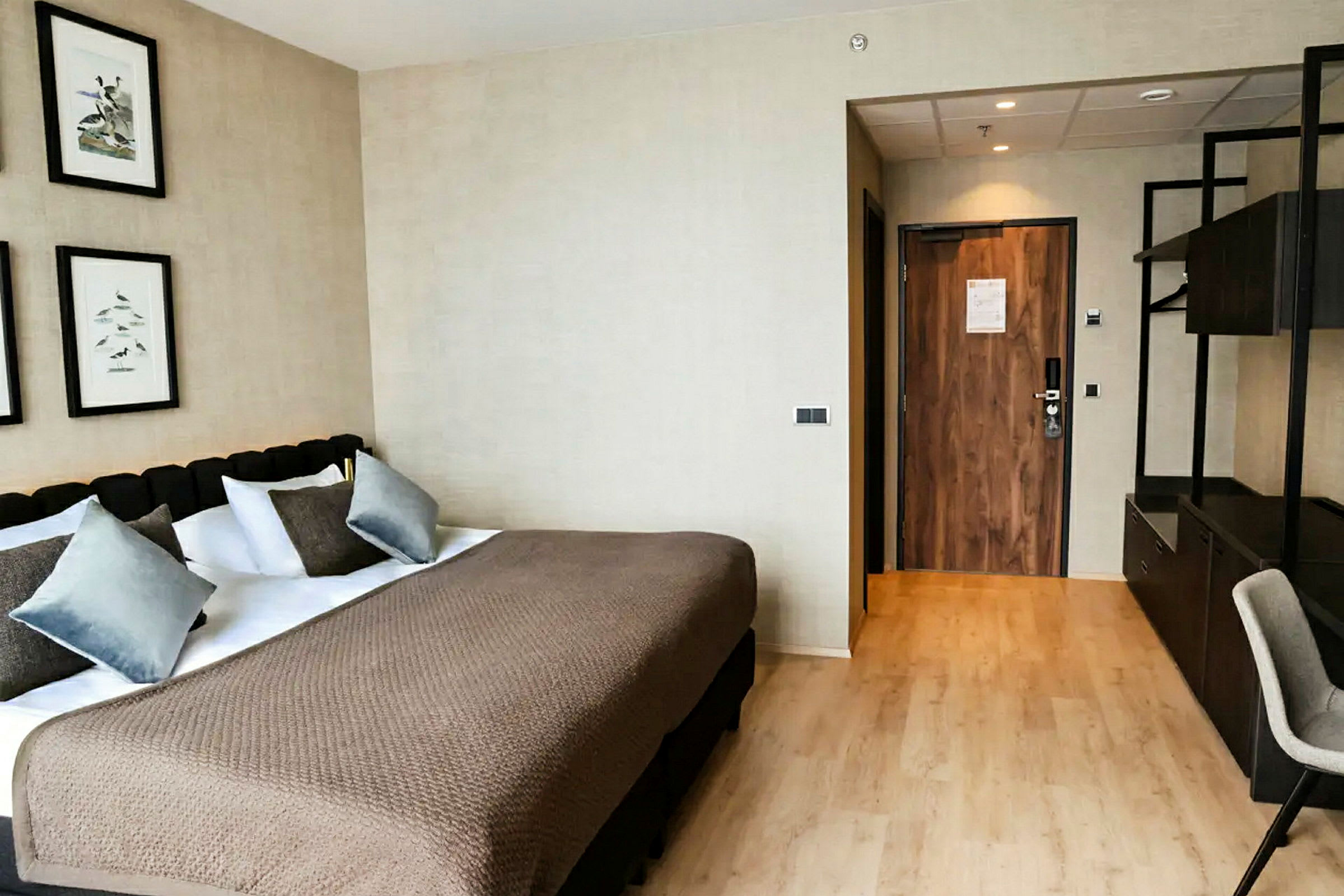 Ett hotellrum med en stor säng, flera kuddar och ett beige överkast. På väggen ovanför hänger tavlor, och en dörr till höger leder ut. (AI-genererad text)