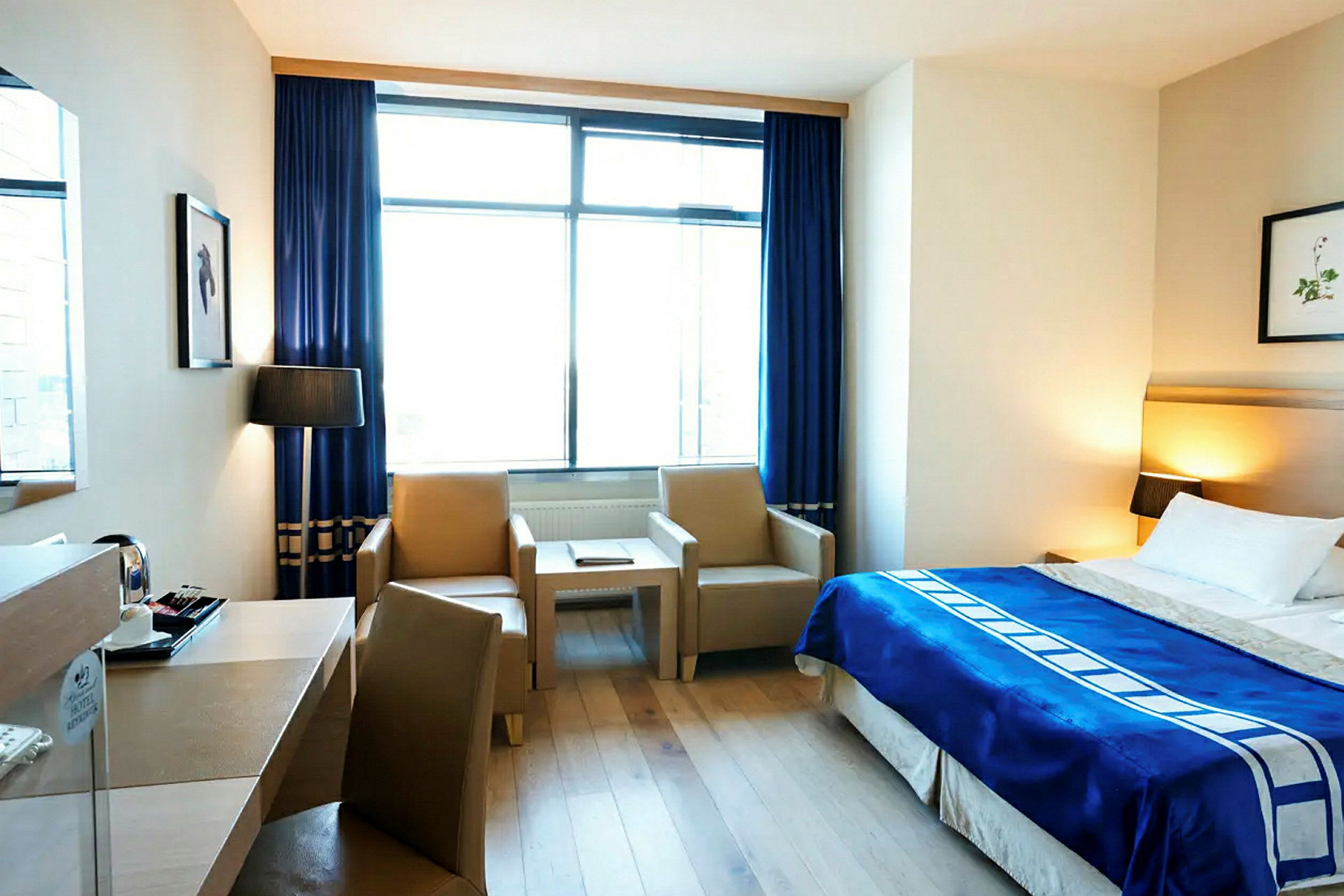 Ett hotellrum med en dubbelsäng, blå täcke och två fåtöljer vid ett soffbord. Ett stort fönster med blå gardiner släpper in dagsljus. (AI-genererad text)