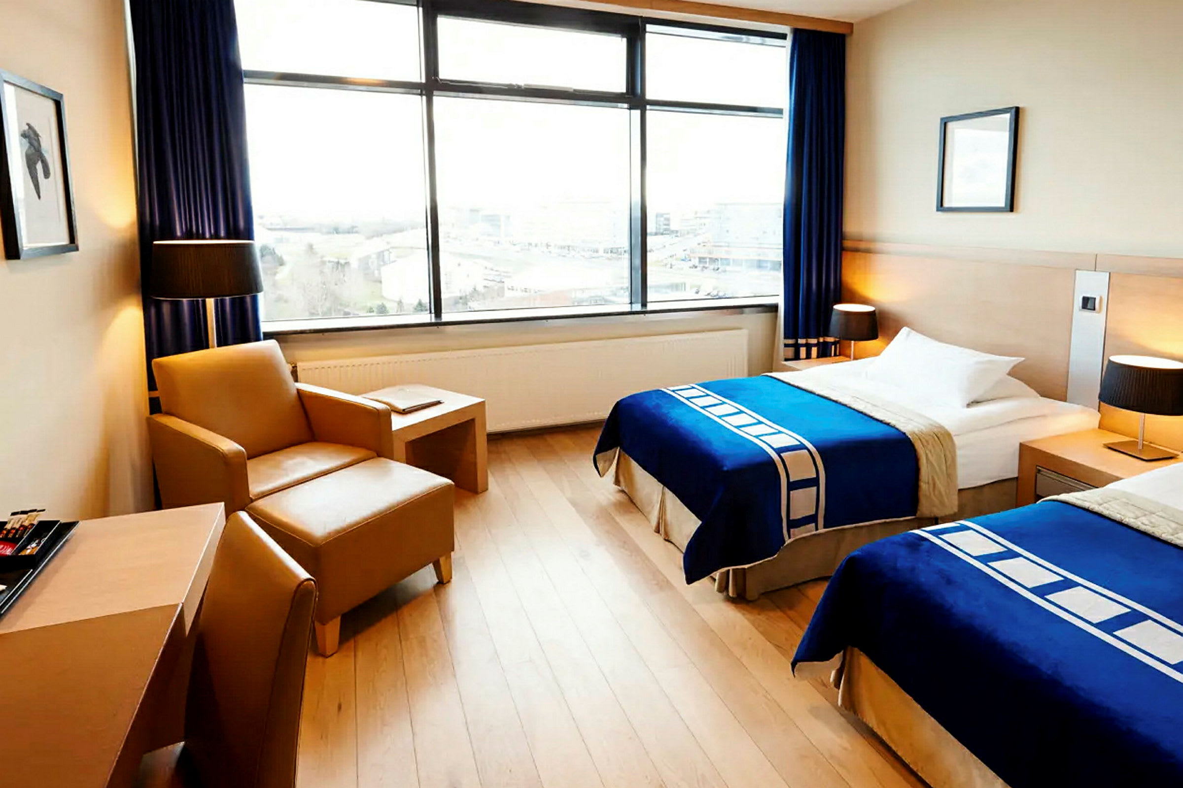 Bild av ett hotellrum med två enkelsängar, blå sängöverkast, en fåtölj med ett bord, och stora fönster med utsikt. (AI-genererad text)