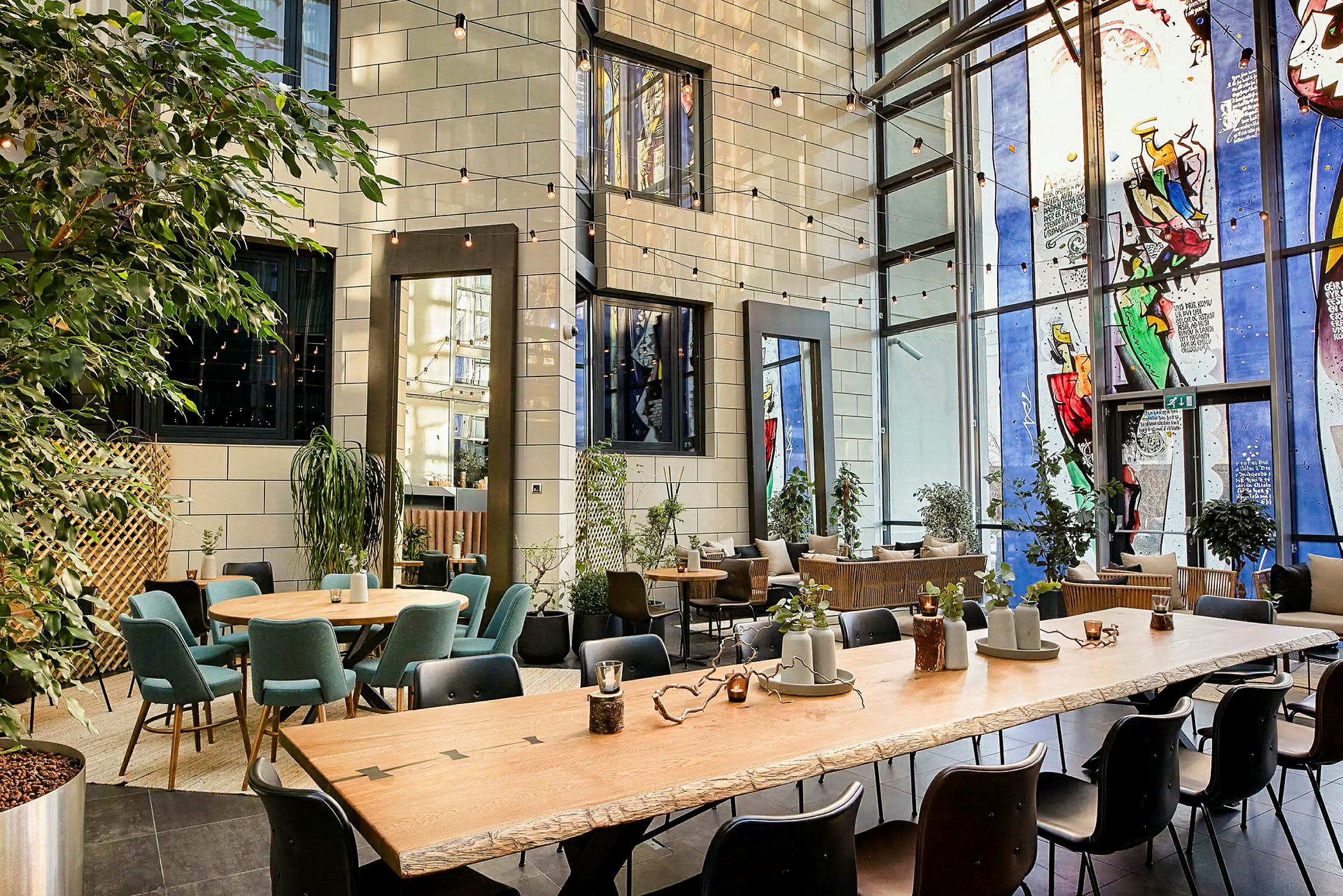 En inomhus lounge med stora fönster, sittplatser runt flera bord och gröna växter placerade i olika områden. (AI-genererad text)