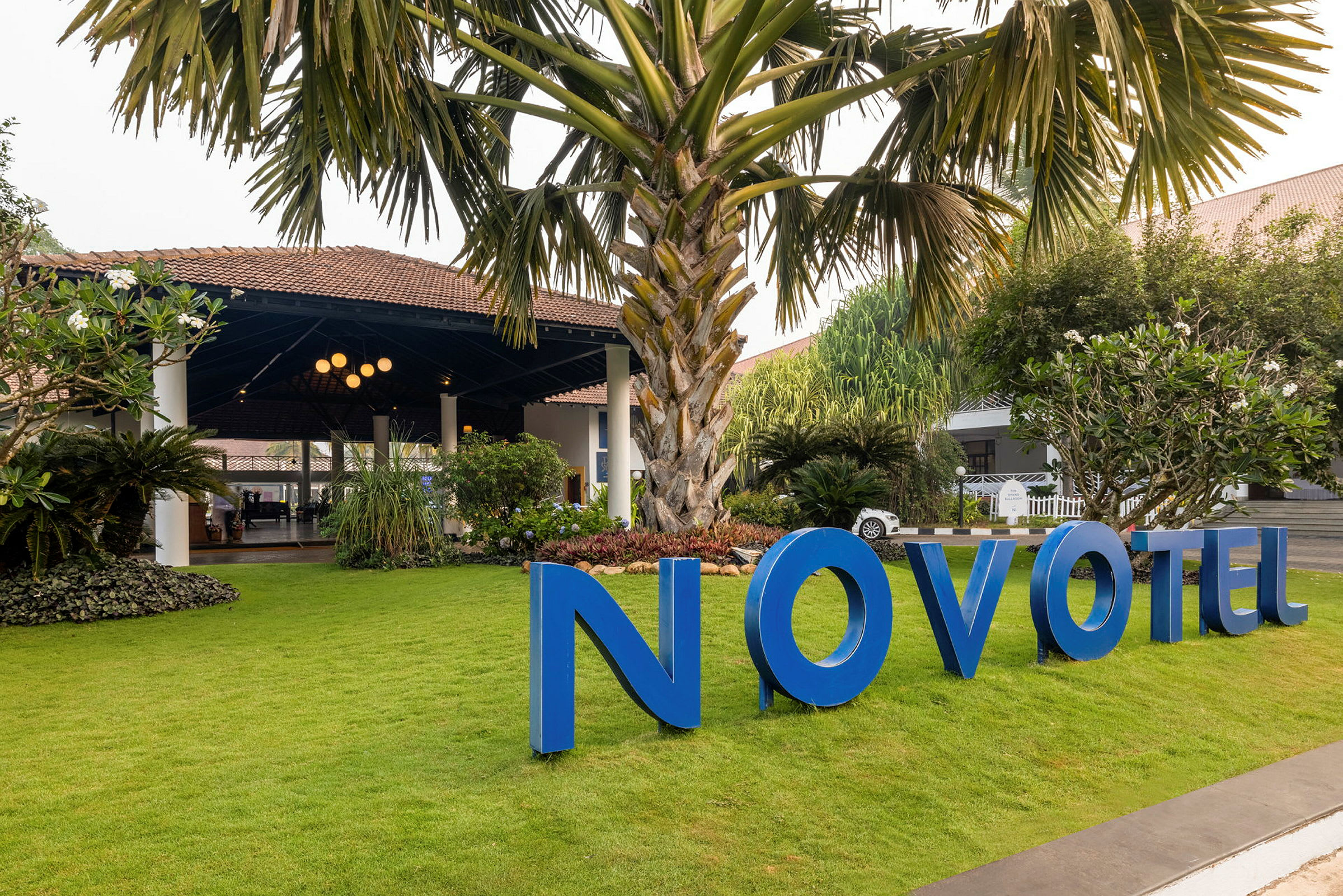 En trädgård med stor palm och grön gräsmatta framför en byggnad med tak och blå skylt som säger "Novotel". (AI-genererad text)