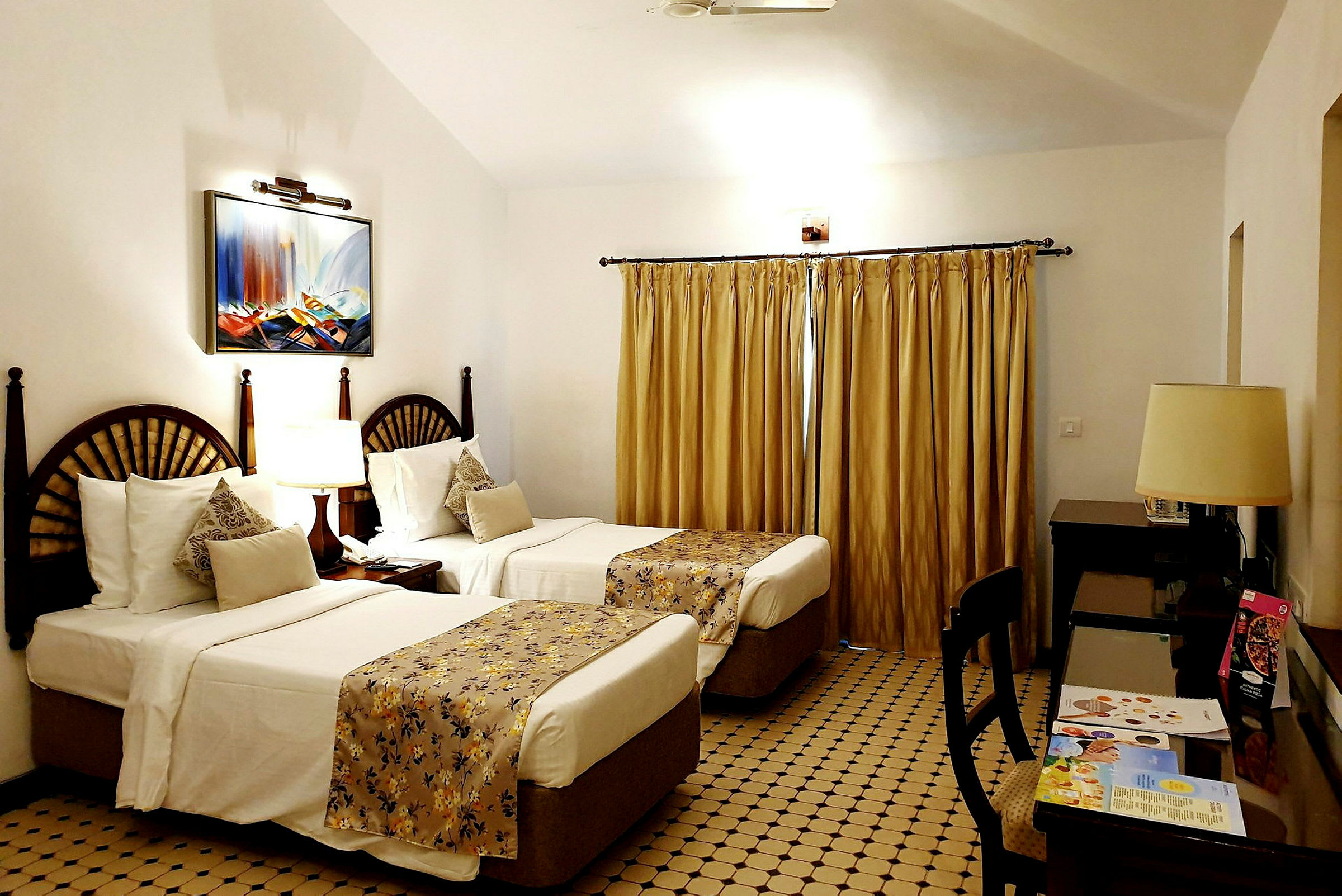Ett hotellrum med två enkelsängar, inredda med vita sängkläder och gulddraperade överkast. Väggarna är vita och fönstret är täckt med gardiner. En tavla pryder väggen ovanför sängarna. (AI-genererad text)