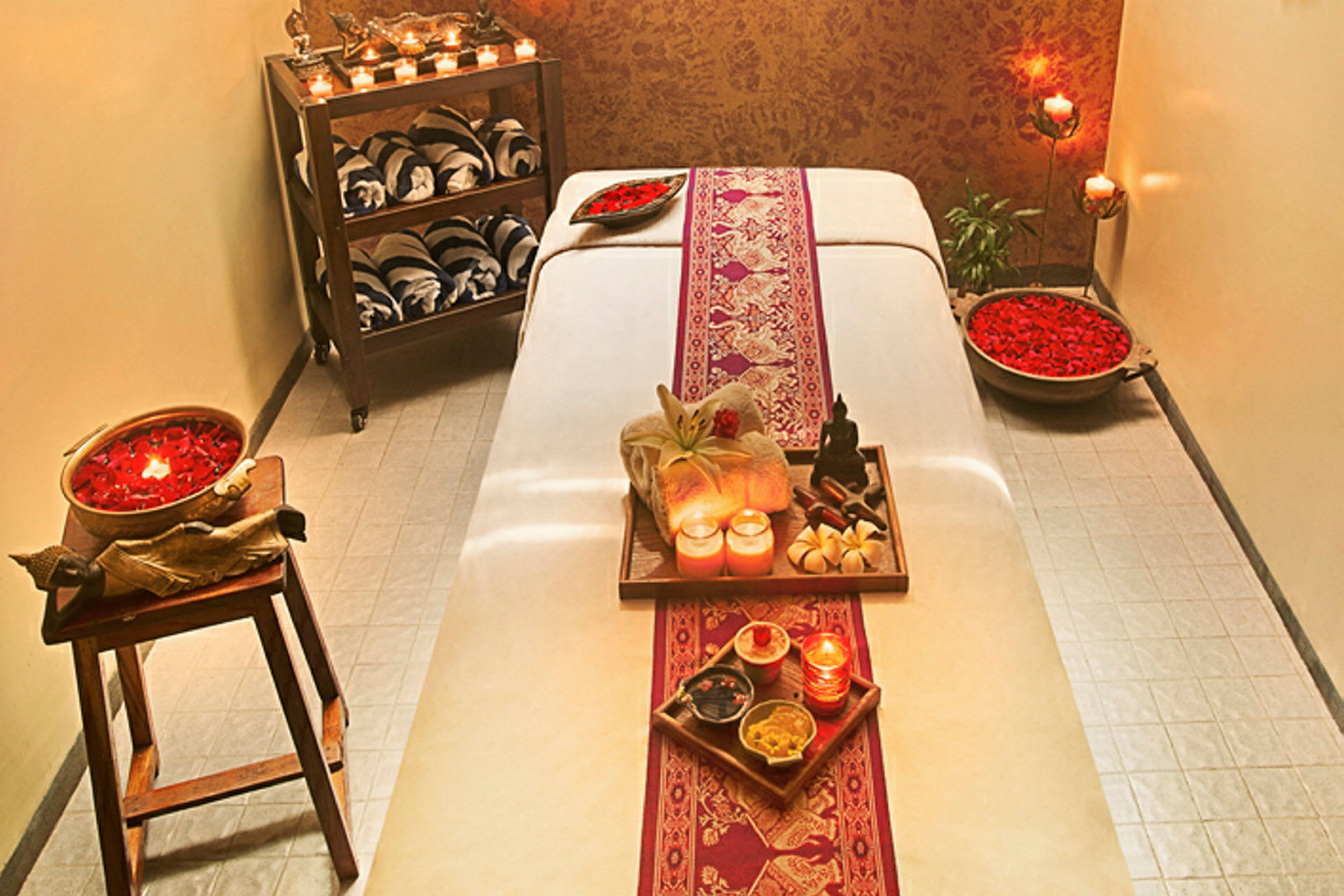 Ett rum inrett för massage med en bänk dekorerad med ljus, blomblad och handdukar. (AI-genererad text)