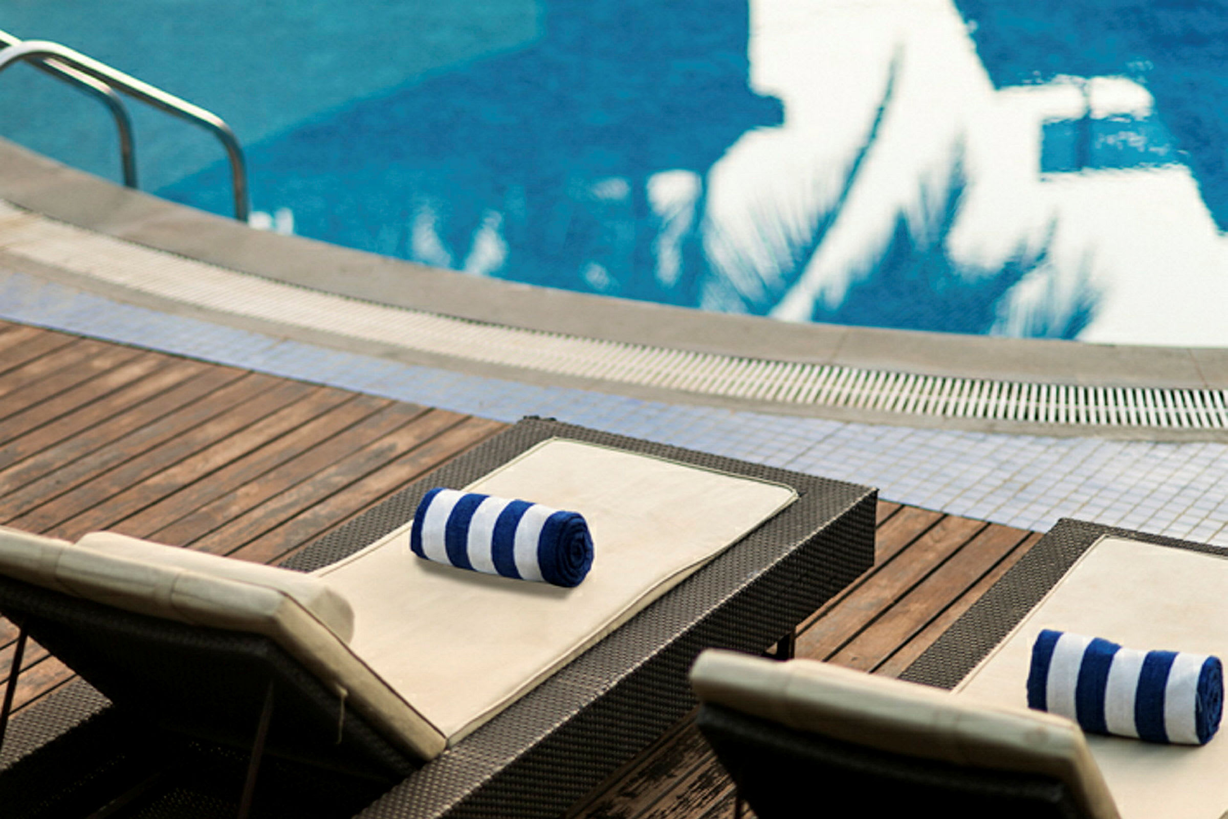 Två solstolar med randiga handdukar ligger på ett trädeck intill en pool. (AI-genererad text)