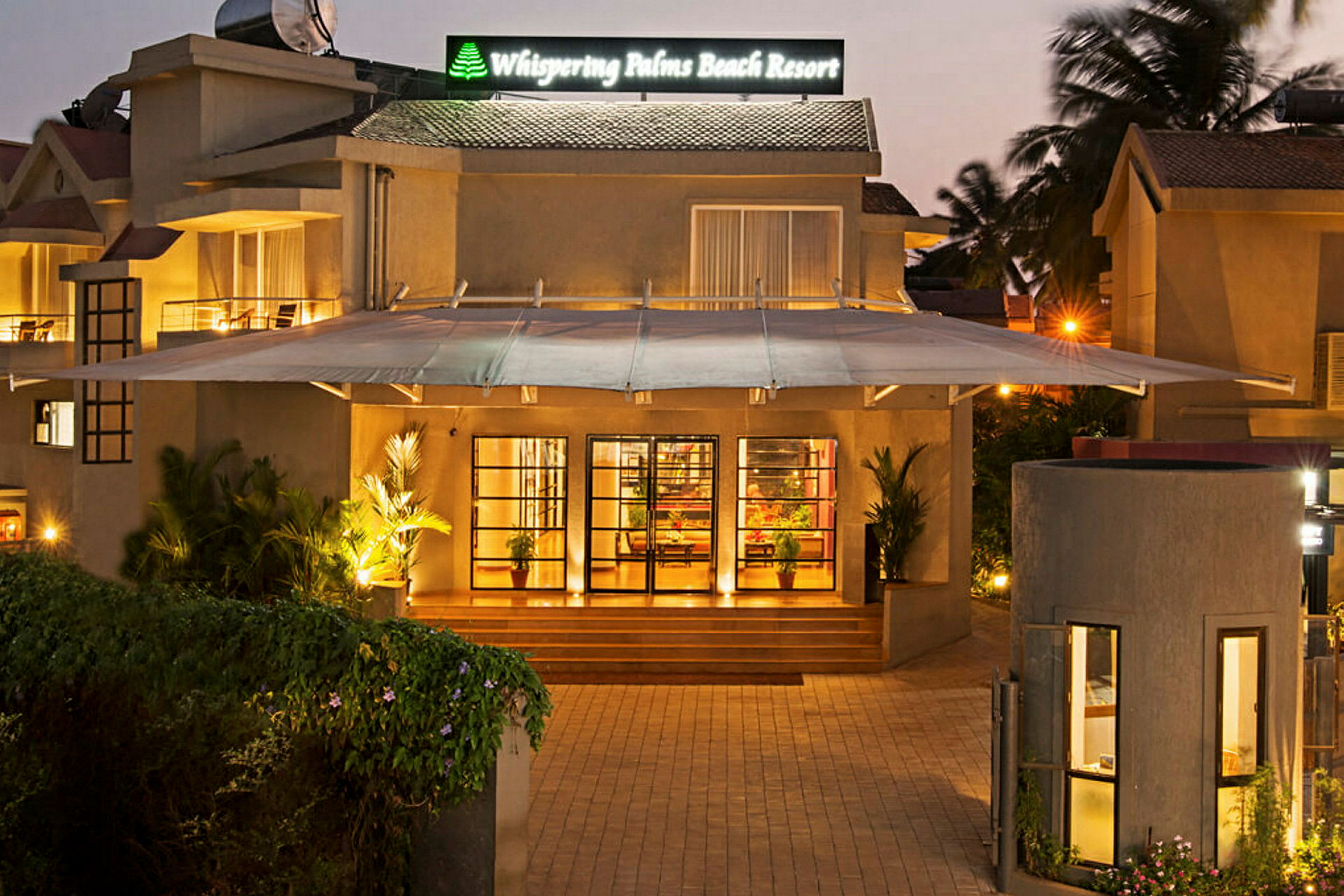 Entréen till Whispering Palms Beach Resort med en upplyst fasad och gröna växter. (AI-genererad text)