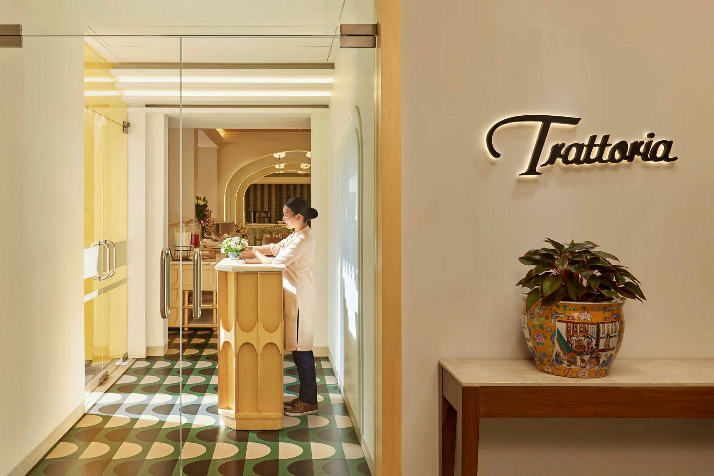 Ingången till en restaurang med ordet 'Trattoria' på väggen och ett bord med krukväxt. (AI-genererad text)
