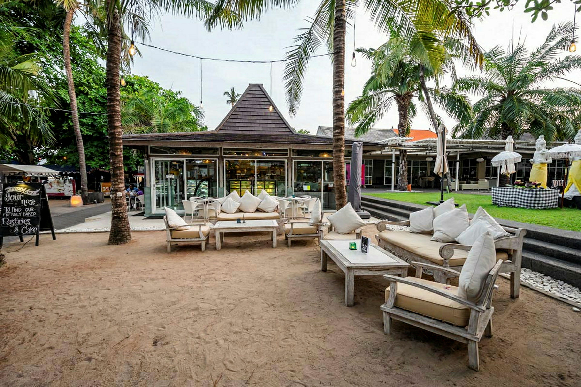 En utomhuslounge med bekväma möbler omgiven av palmer och ett café eller restaurang i bakgrunden. (AI-genererad text)