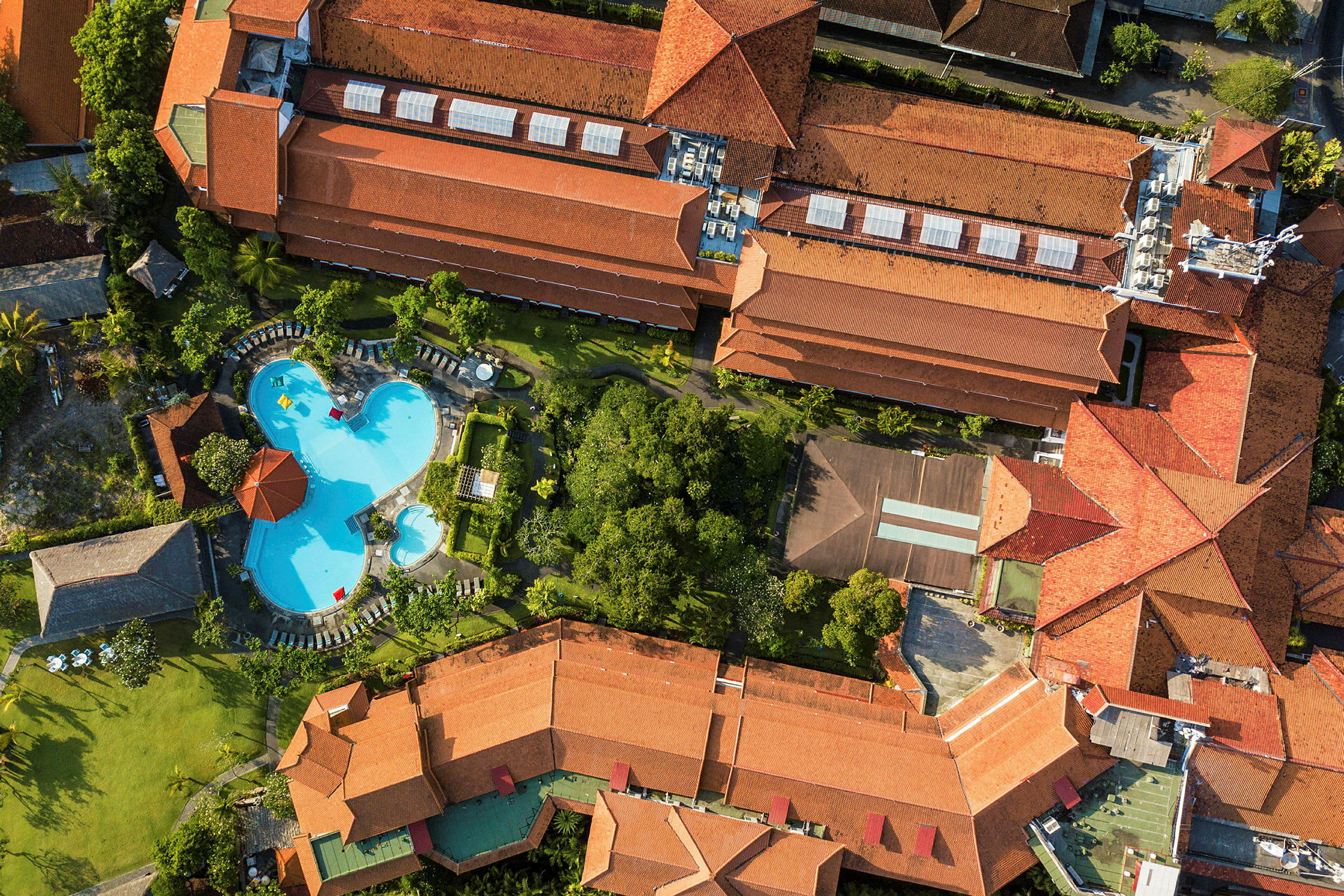 Flygbild av en resort med en stor pool, grönområden och byggnader med röda tak. (AI-genererad text)