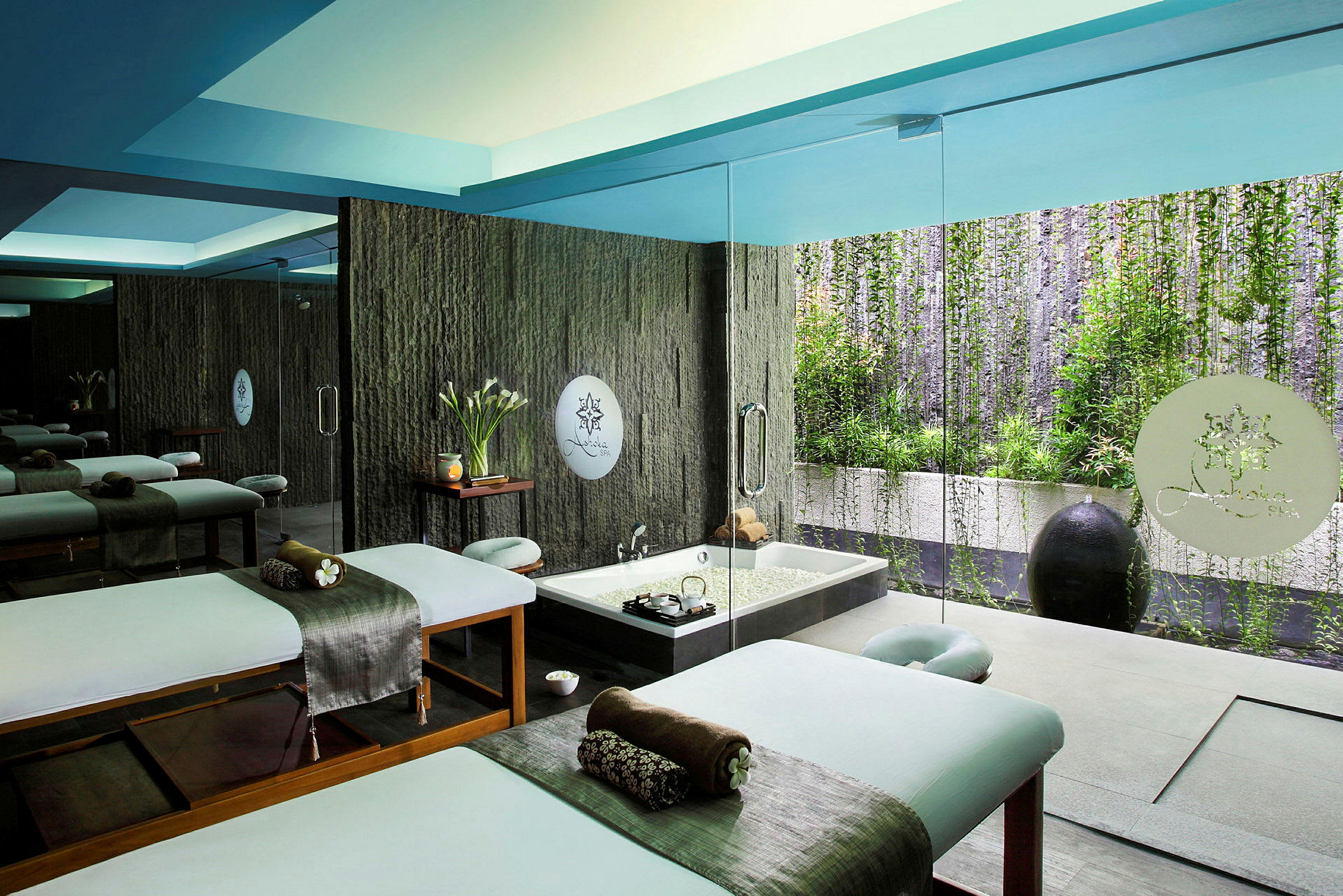 Ett spaområde med massagebänkar och en jacuzzi, omgiven av glasväggar och grönska. (AI-genererad text)
