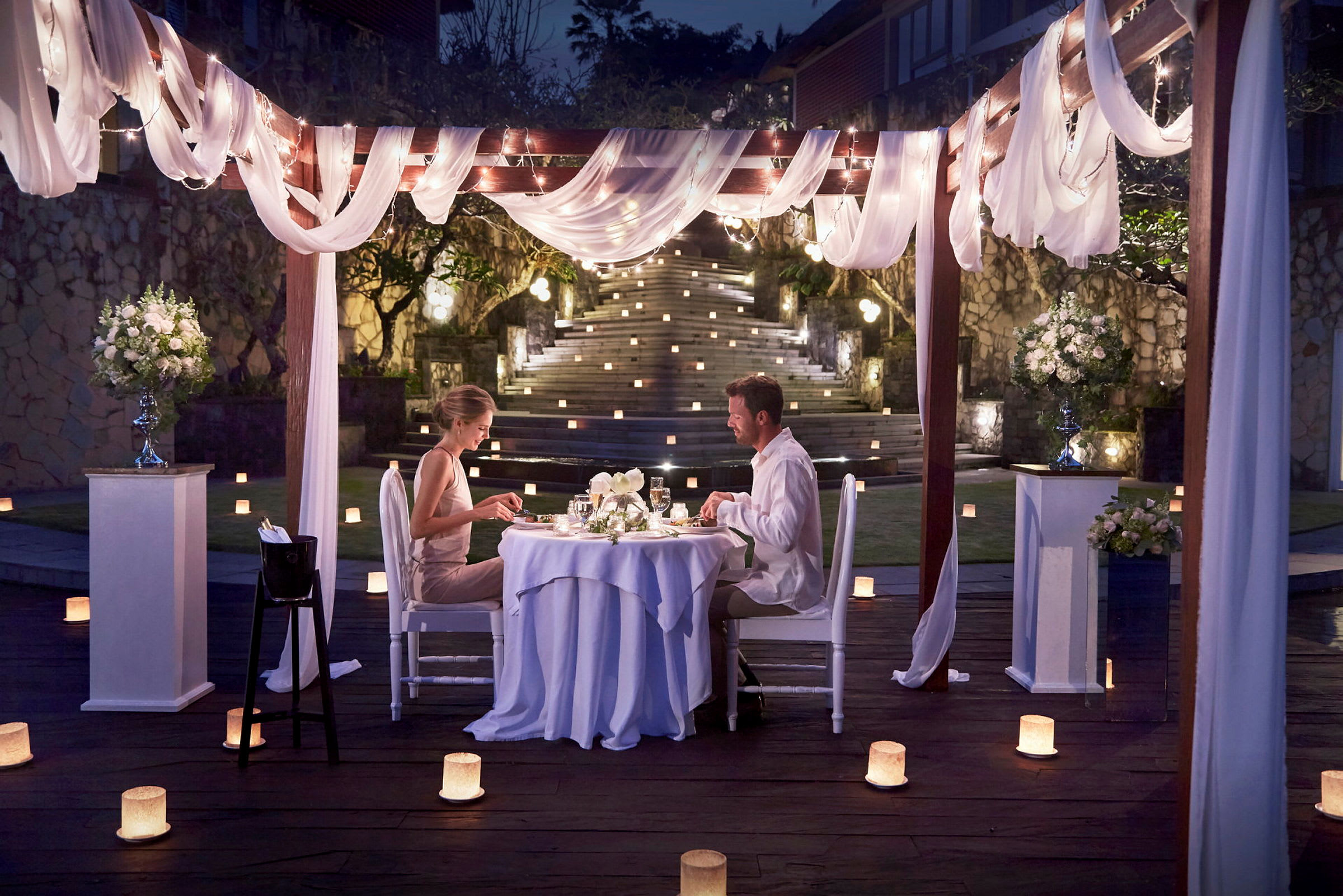 Ett middagsbord utomhus under en elegant dekorerad pergola med ljus och blommor. (AI-genererad text)