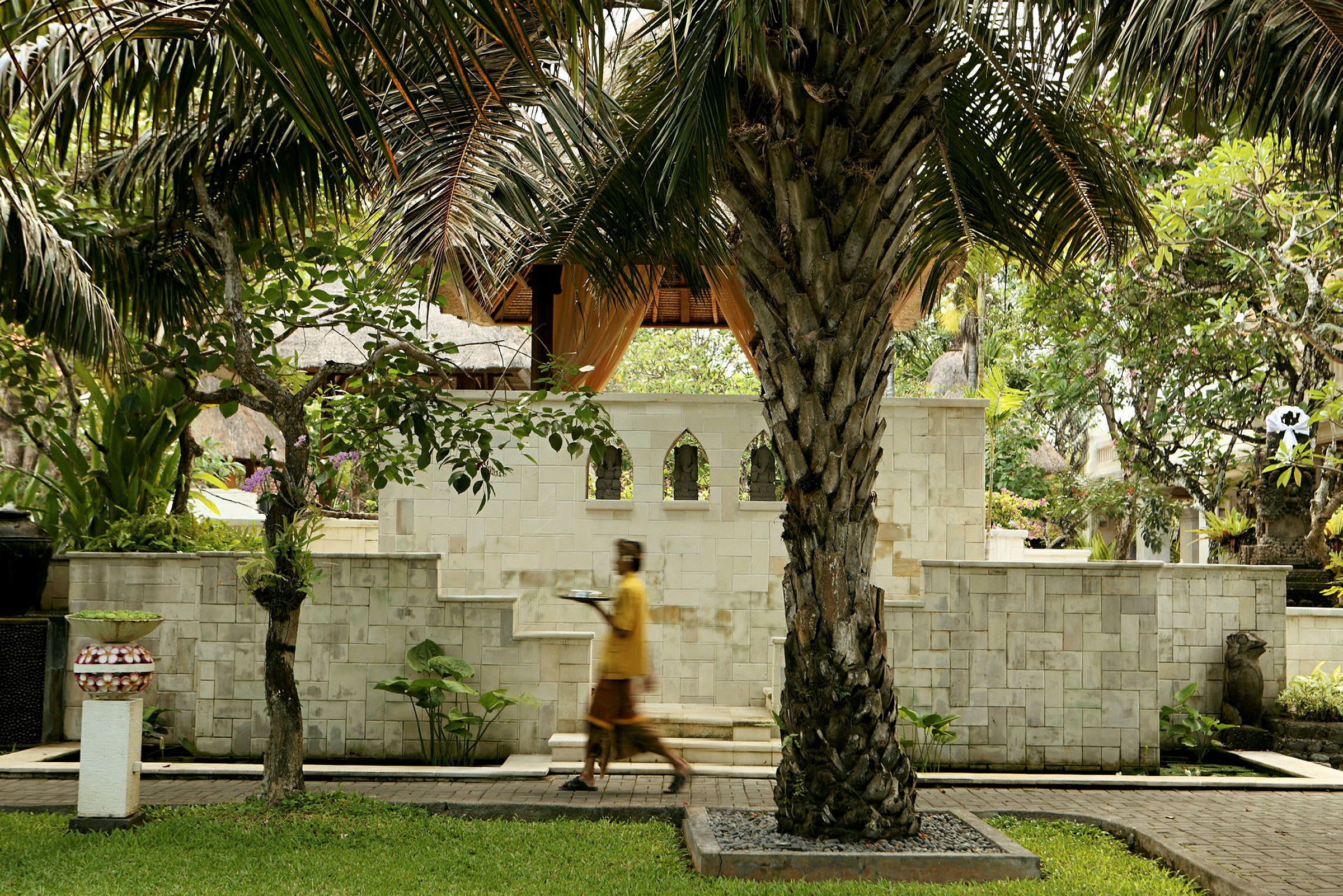 En person i gul dräkt går förbi en trädgård med palmer och stenbänkar. (AI-genererad text)