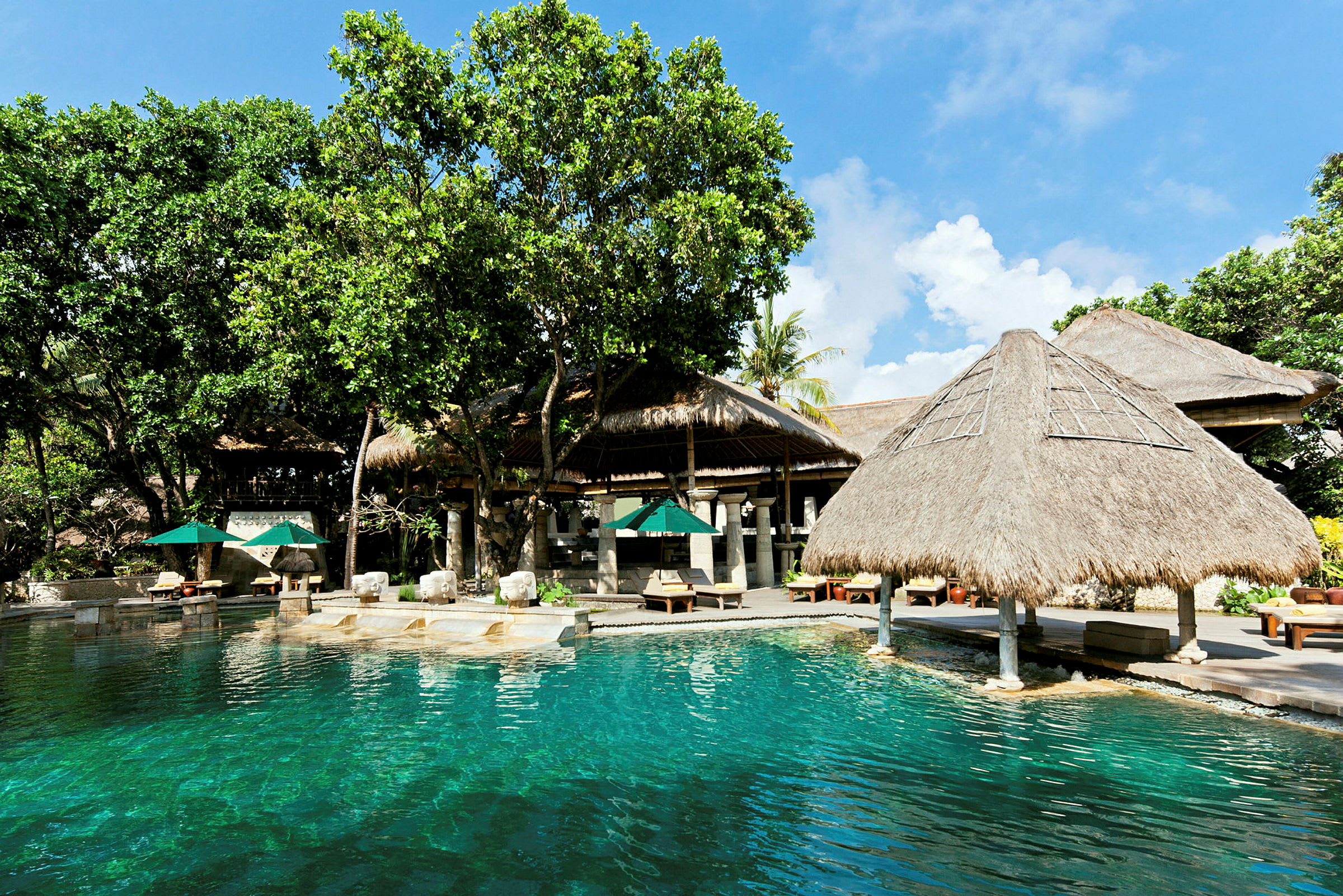 En utomhuspool med klart vatten omgivet av träd och halmtakade strukturer. Solparasoller och solstolar är placerade på terrassen intill. (AI-genererad text)