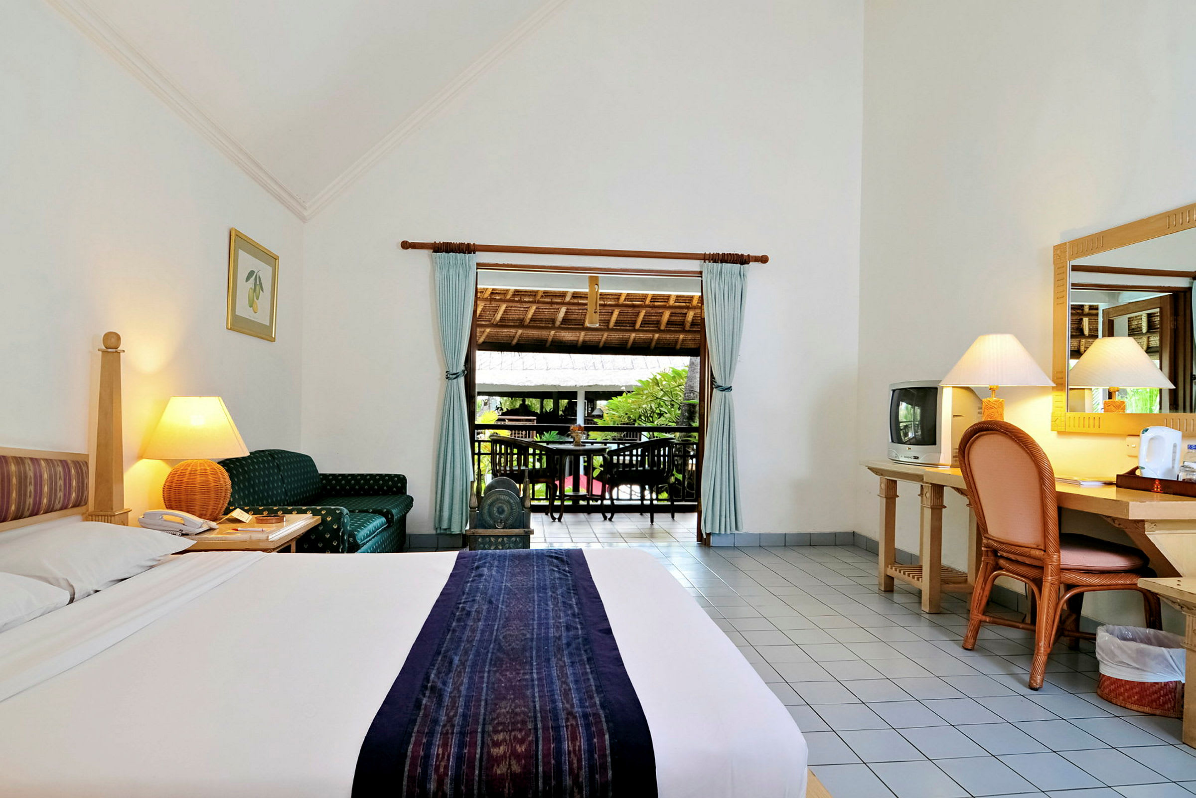 Ett hotellrum med en säng, skrivbord, stol och en öppning till en balkong med utsikt mot grönska. (AI-genererad text)