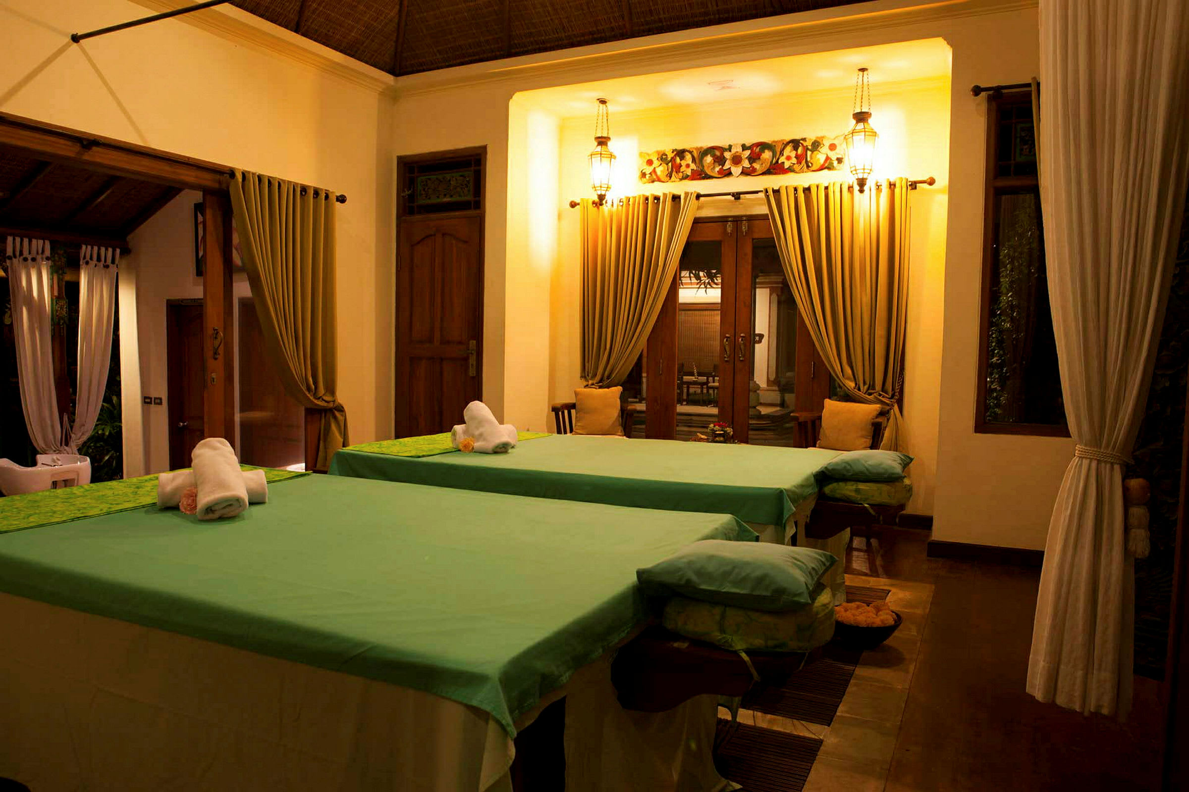 Två massagebänkar med gröna överdrag i ett upplyst spa-rum med draperier och träinredning. (AI-genererad text)