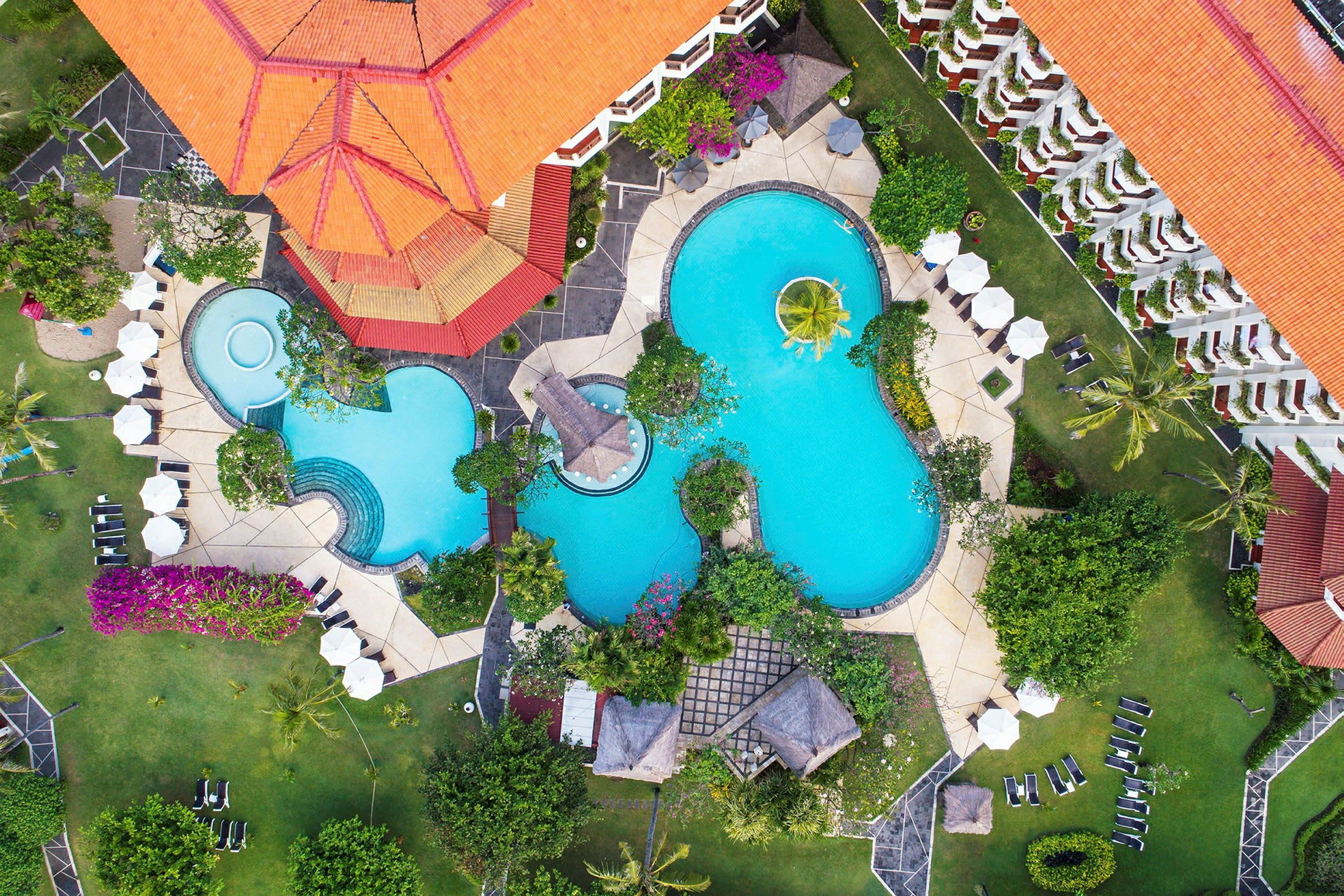 Flygbild på en resort med flera pooler, omgärdade av gröna trädgårdar och solstolar. (AI-genererad text)