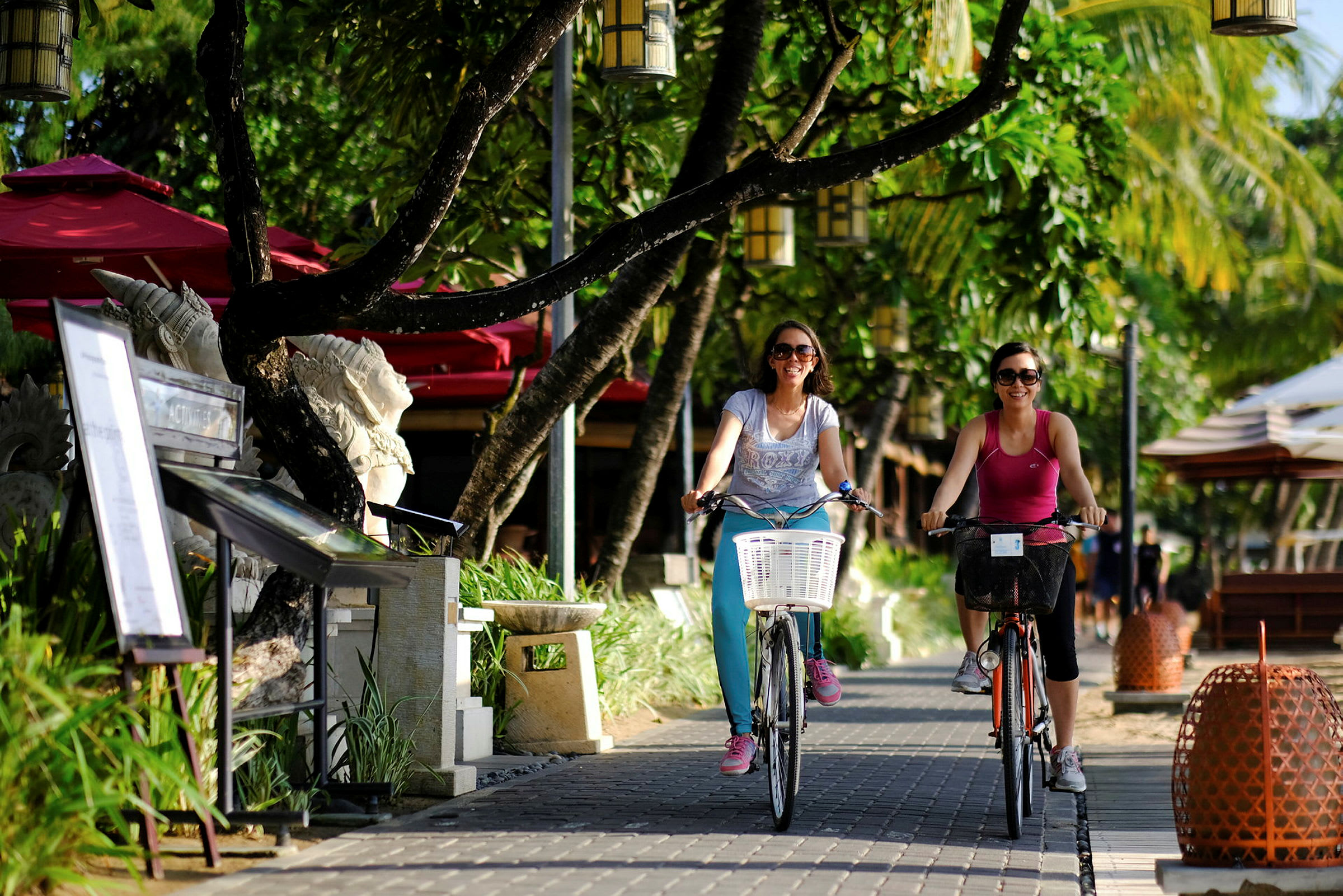 Två personer cyklar längs en stenlagd gångväg omgiven av gröna träd och lampor. (AI-genererad text)