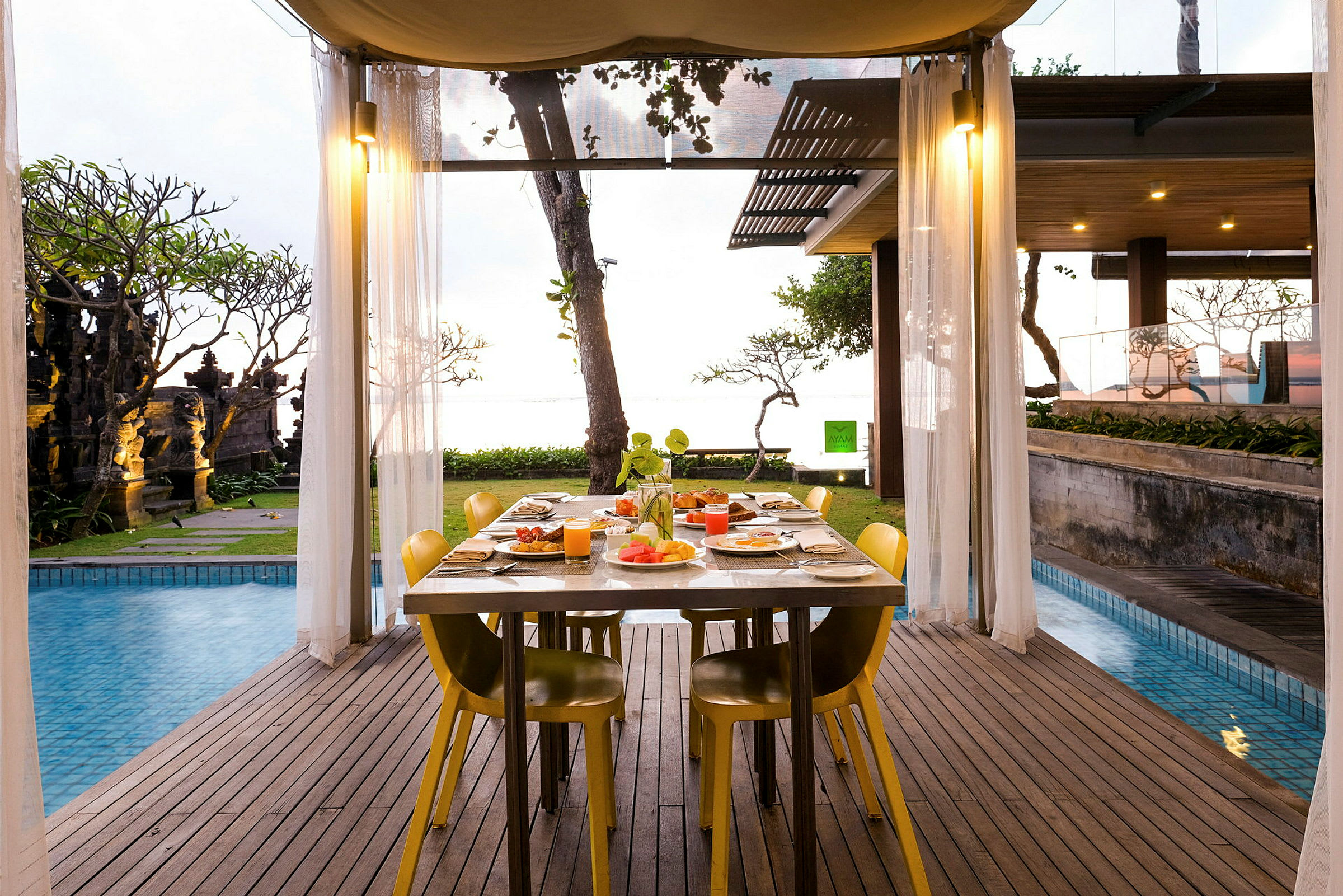 Ett bord dukat för måltid står under en övertäckt terrass nära en pool med utsikt mot trädgård och hav. (AI-genererad text)