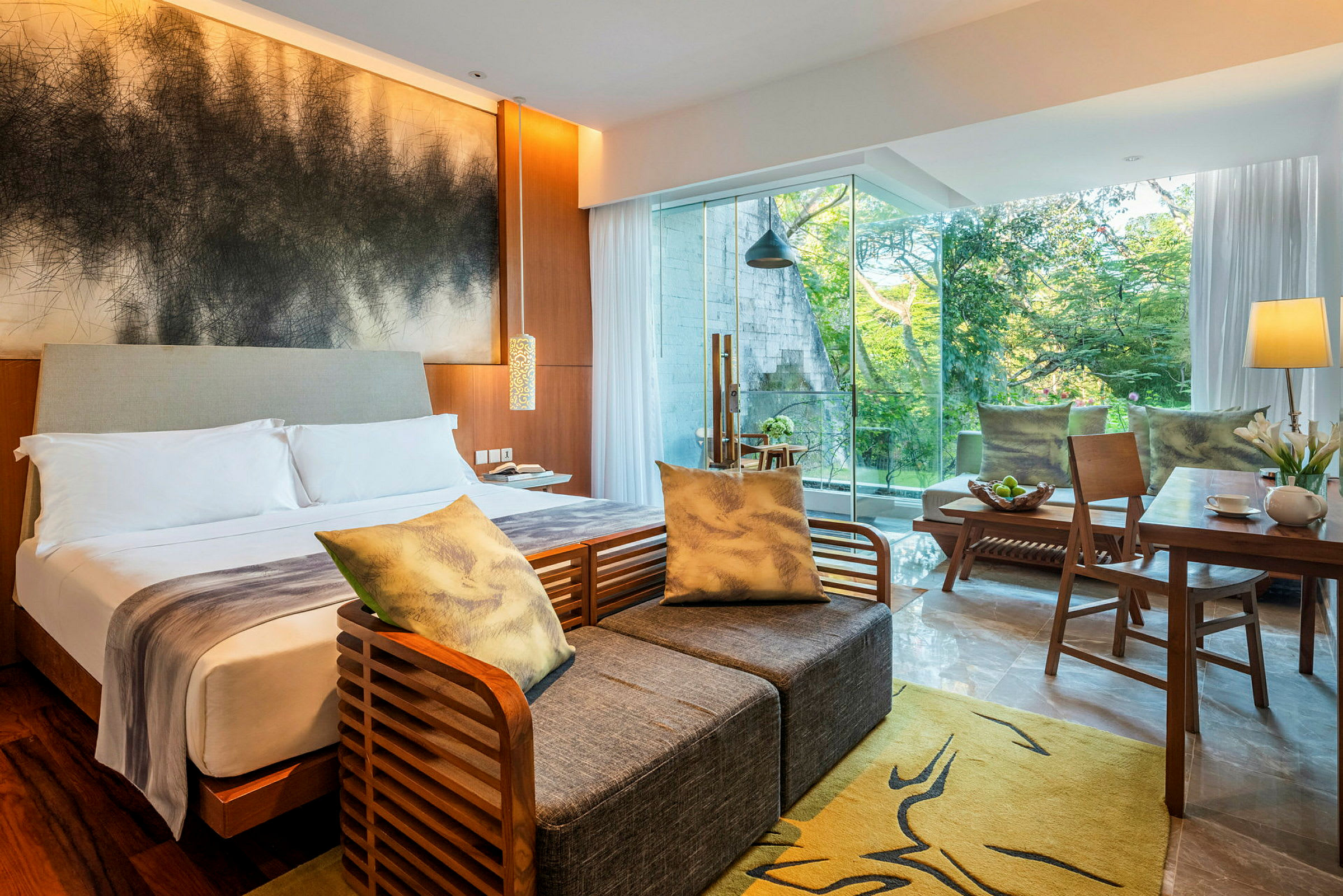 Ett modernt hotellrum med en dubbelsäng, soffmöbler, ett skrivbord och stora fönster som visar grönska utanför. (AI-genererad text)