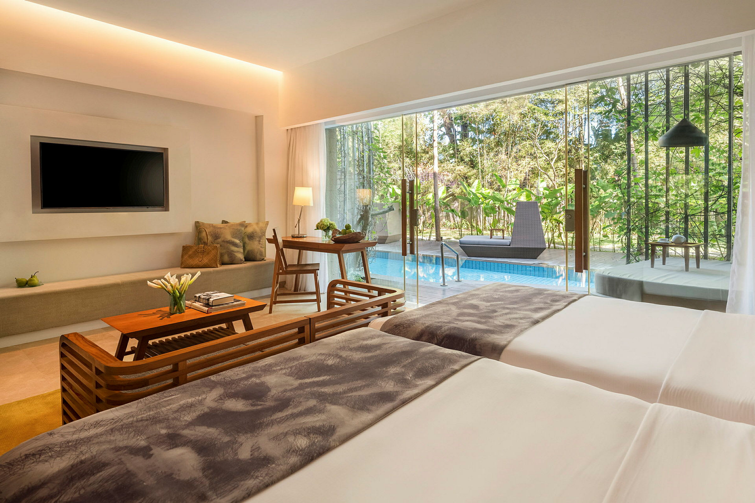 Ett modernt hotellrum med sängar, soffhörna, TV, matbord och utsikt mot en pool och grönska utanför glasdörrar. (AI-genererad text)