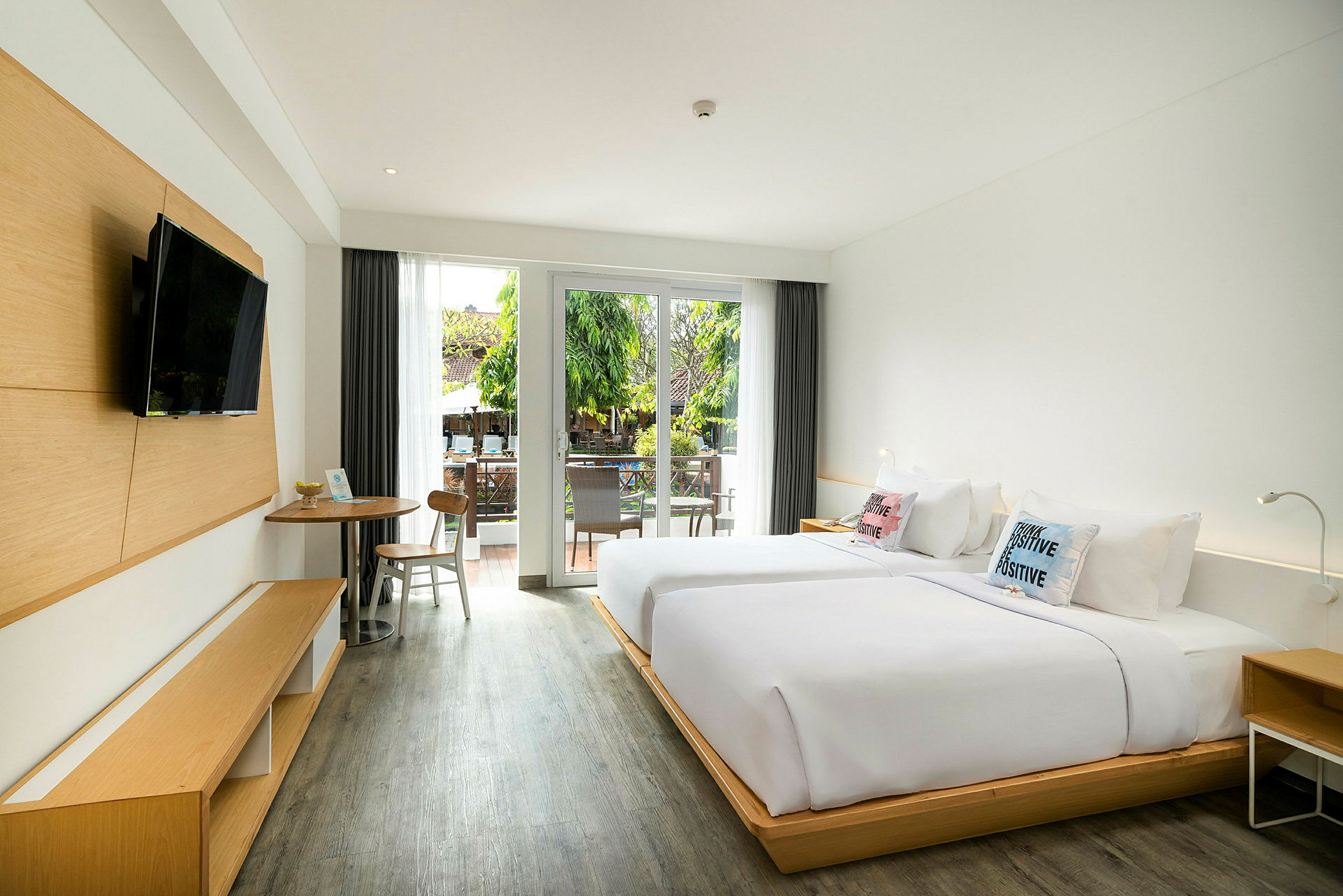 Ett hotellrum med två sängar, ett bord med stolar och en modern TV på väggen. Rummet har stora glasdörrar som leder ut till en balkong med utsikt över grönska. (AI-genererad text)