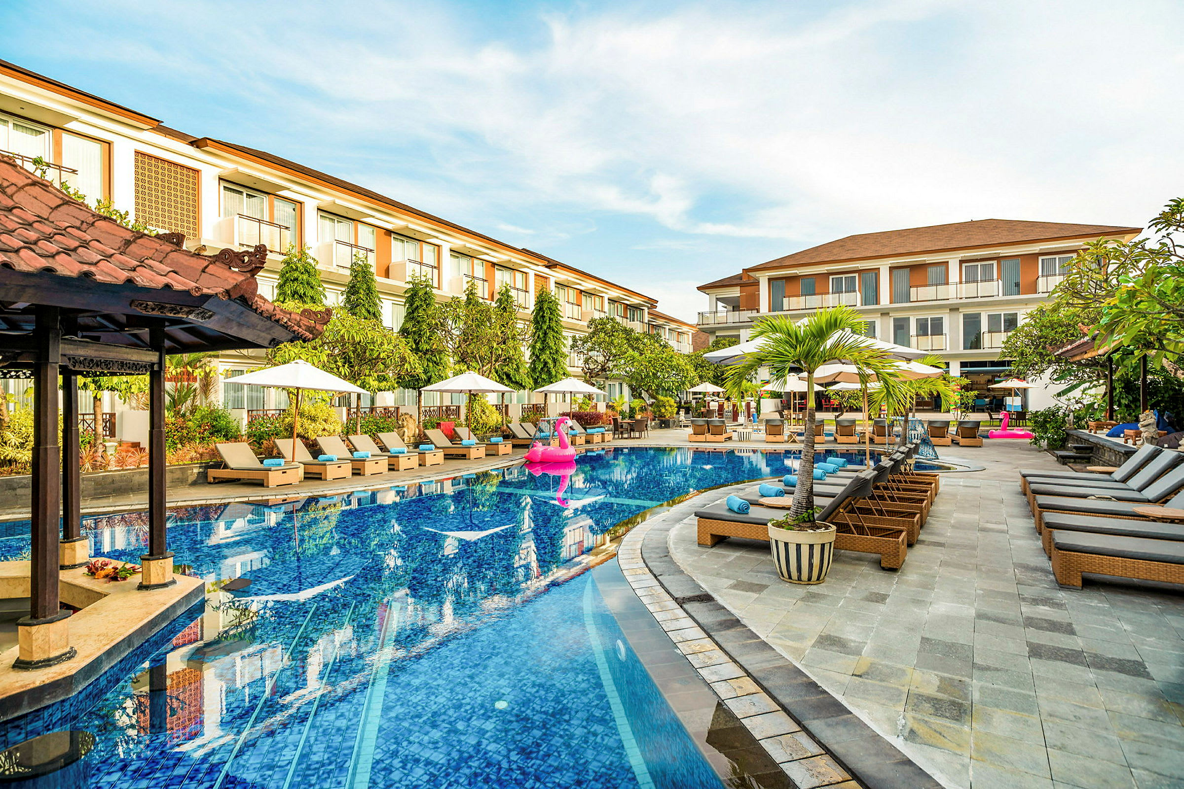 En hotellpool med solsängar och parasoller runt omkring, samt ett par byggnader i bakgrunden. (AI-genererad text)