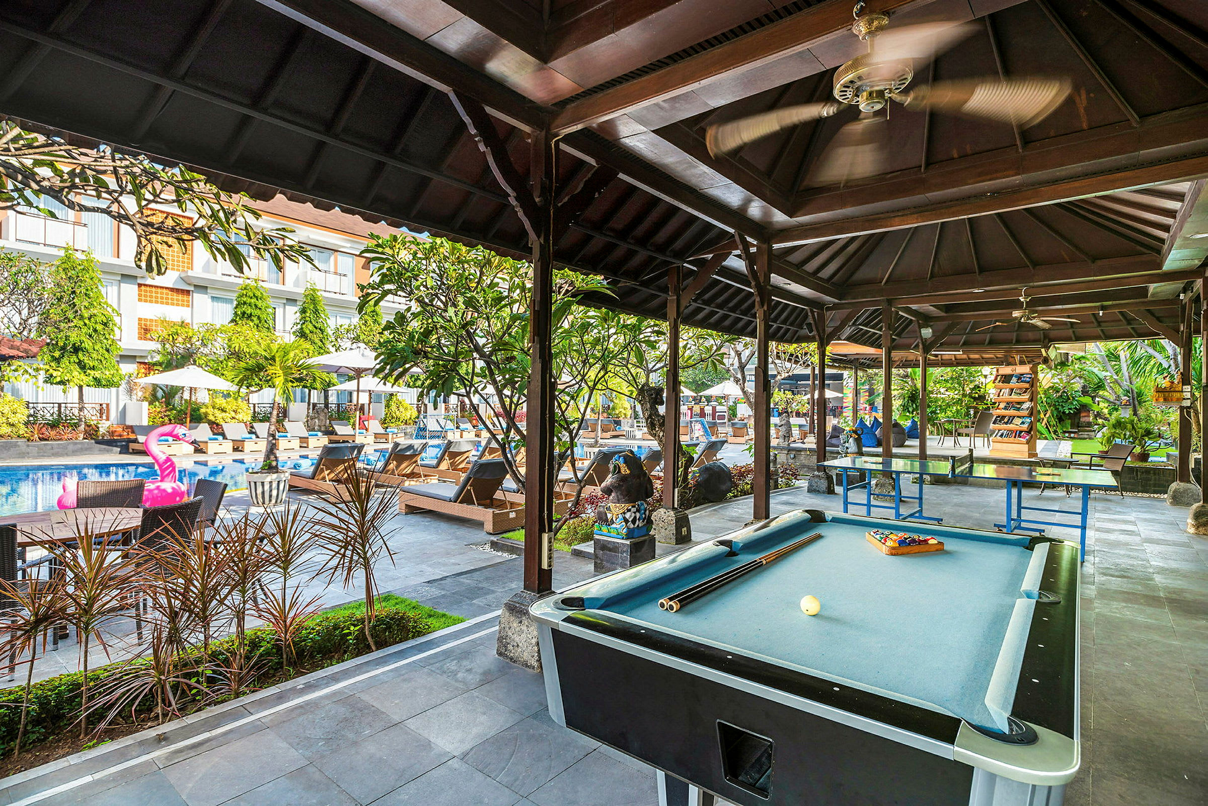 Ett utrymme under tak med biljardbord och pingisbord. I bakgrunden syns en pool med solstolar och parasoller samt en uppblåsbar flamingo. (AI-genererad text)