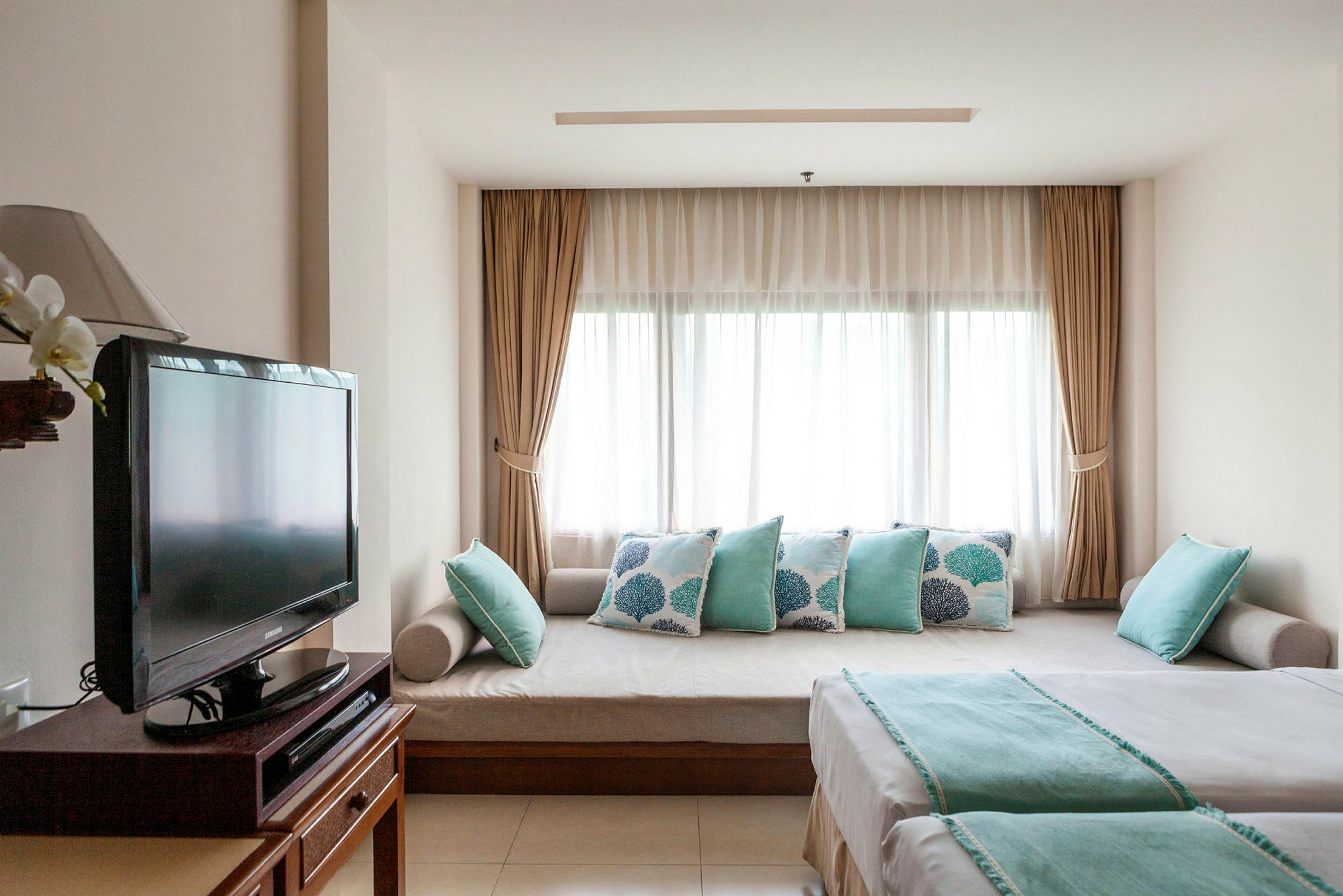 Ett hotellrum med en soffa och kuddar vid ett fönster, en TV och en säng med turkosa detaljer. (AI-genererad text)