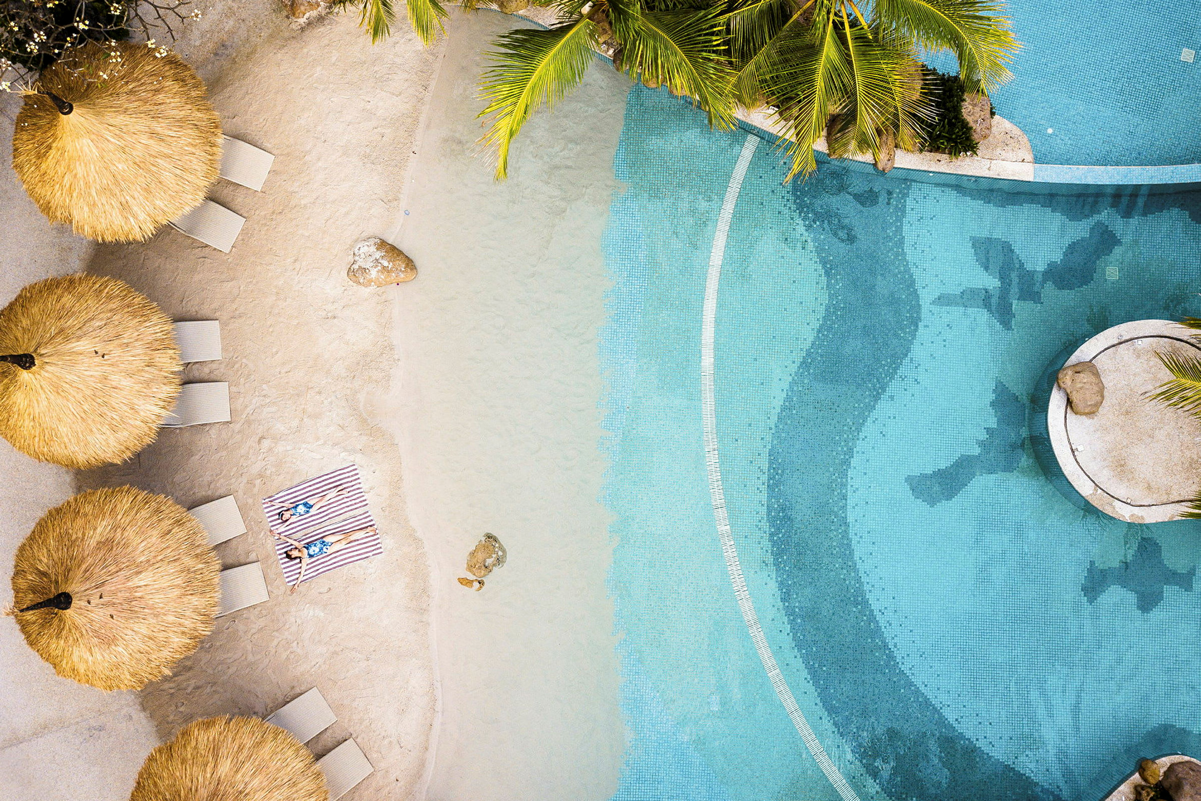 En strand med solstolar placerade under halmtaksskydd vid en pool. (AI-genererad text)