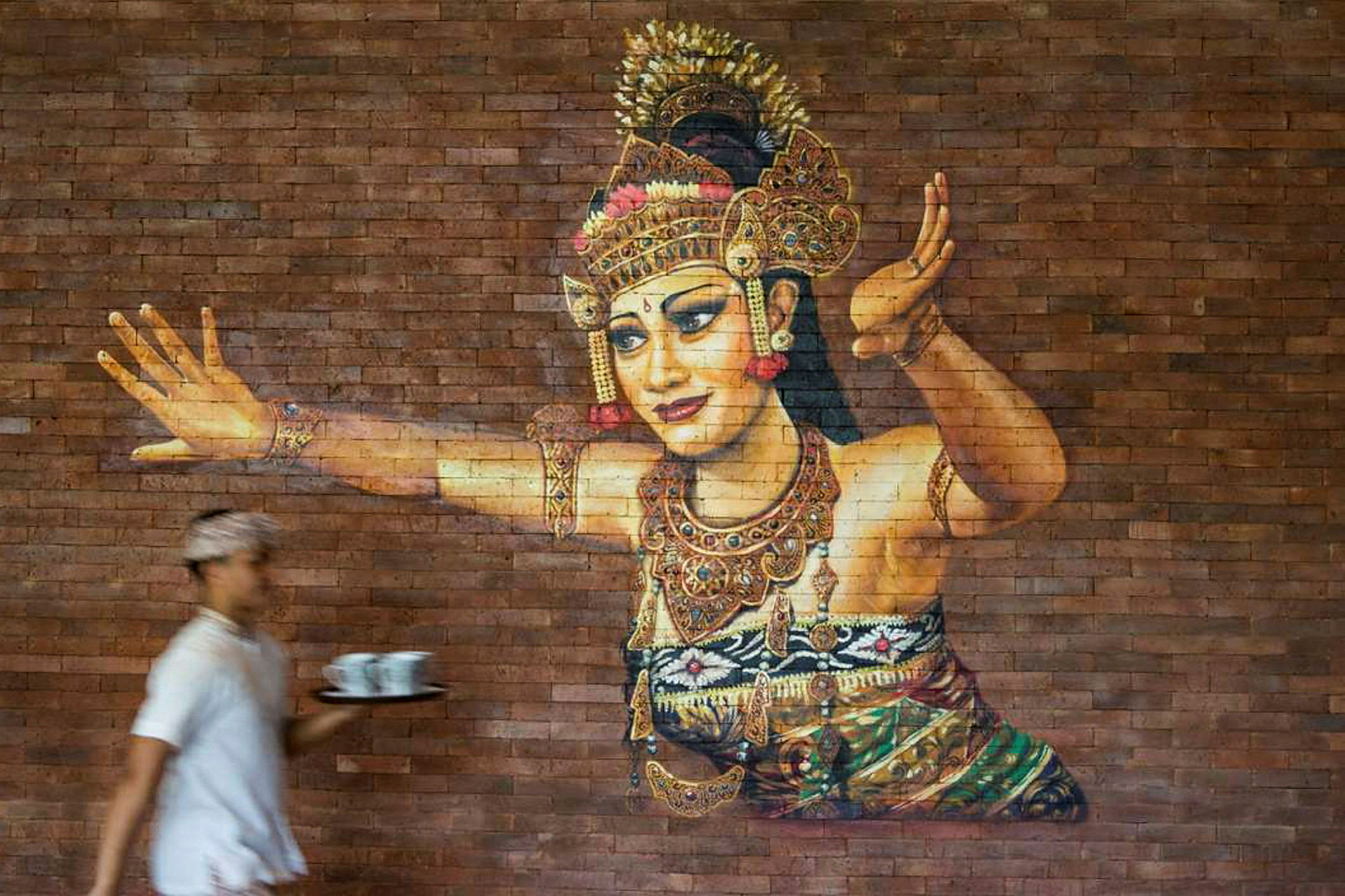 Ett konstverk på en tegelvägg som föreställer en traditionell dansare i utsmyckade kläder och smycken. En person i förgrunden håller ett bricka med koppar. (AI-genererad text)