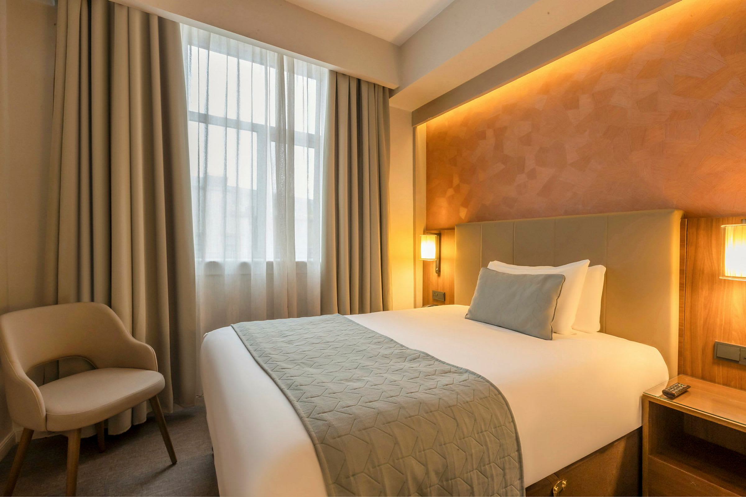 Ett hotellrum med en säng, en stol och ett fönster med gardiner. (AI-genererad text)