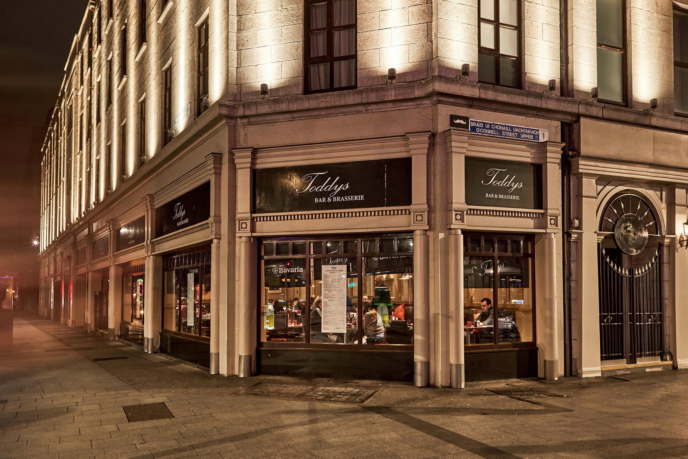 En byggnad av sten med belysta fasader och en restaurang markerad med skyltar som visar Toddy's Bar & Brasserie. (AI-genererad text)