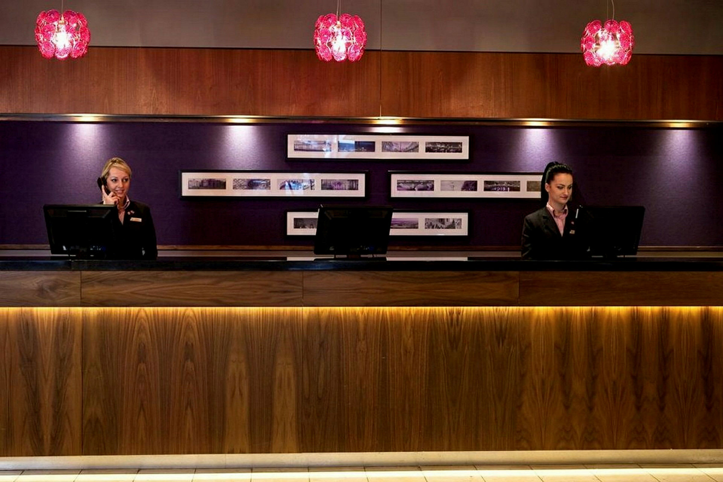 Två receptionister står bakom en receptionsdisk, med datorskärmar och telefoner. Bakgrunden har en klädd vägg med inramade bilder. (AI-genererad text)